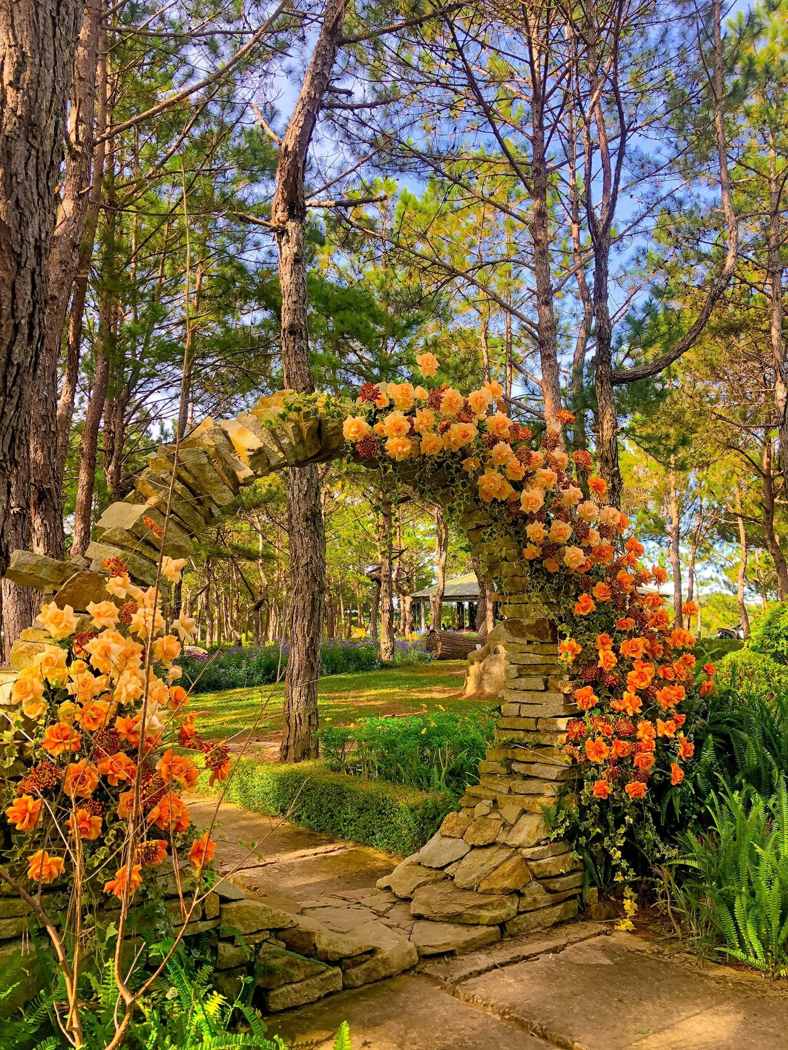 A garden scene with a stone archway decorated with yellow and orange flowers, surrounded by trees and greenery.