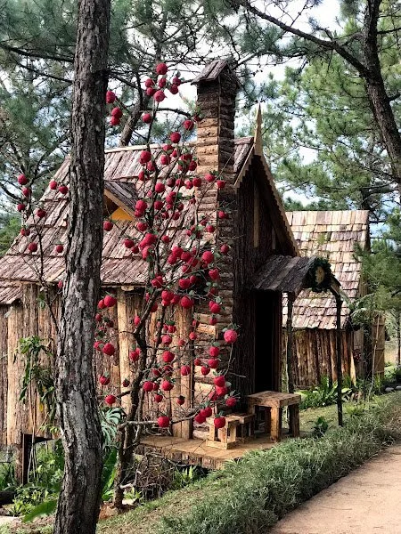 A rustic wooden house with a chimney, surrounded by trees, with a branch of red apples in the foreground.