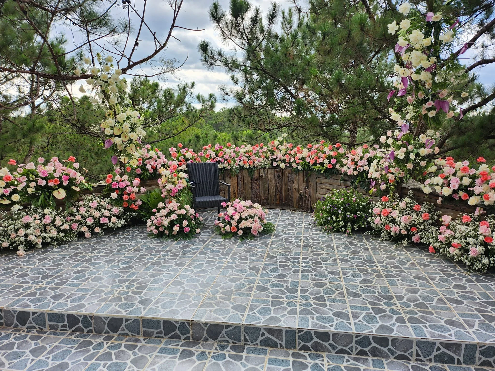 A decorated outdoor platform with a black chair, surrounded by pink and white flowers and greenery, under a cloudy sky.