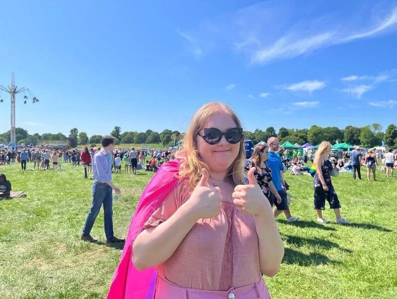 A picture of Emily at pride. she is a white woman with blonde hair, and is wearing all pink with a bisexual flag over her shoulders like a cape. She has sunglasses on and is smiling with two thumbs up. Crowds are behind her with festival rides.
