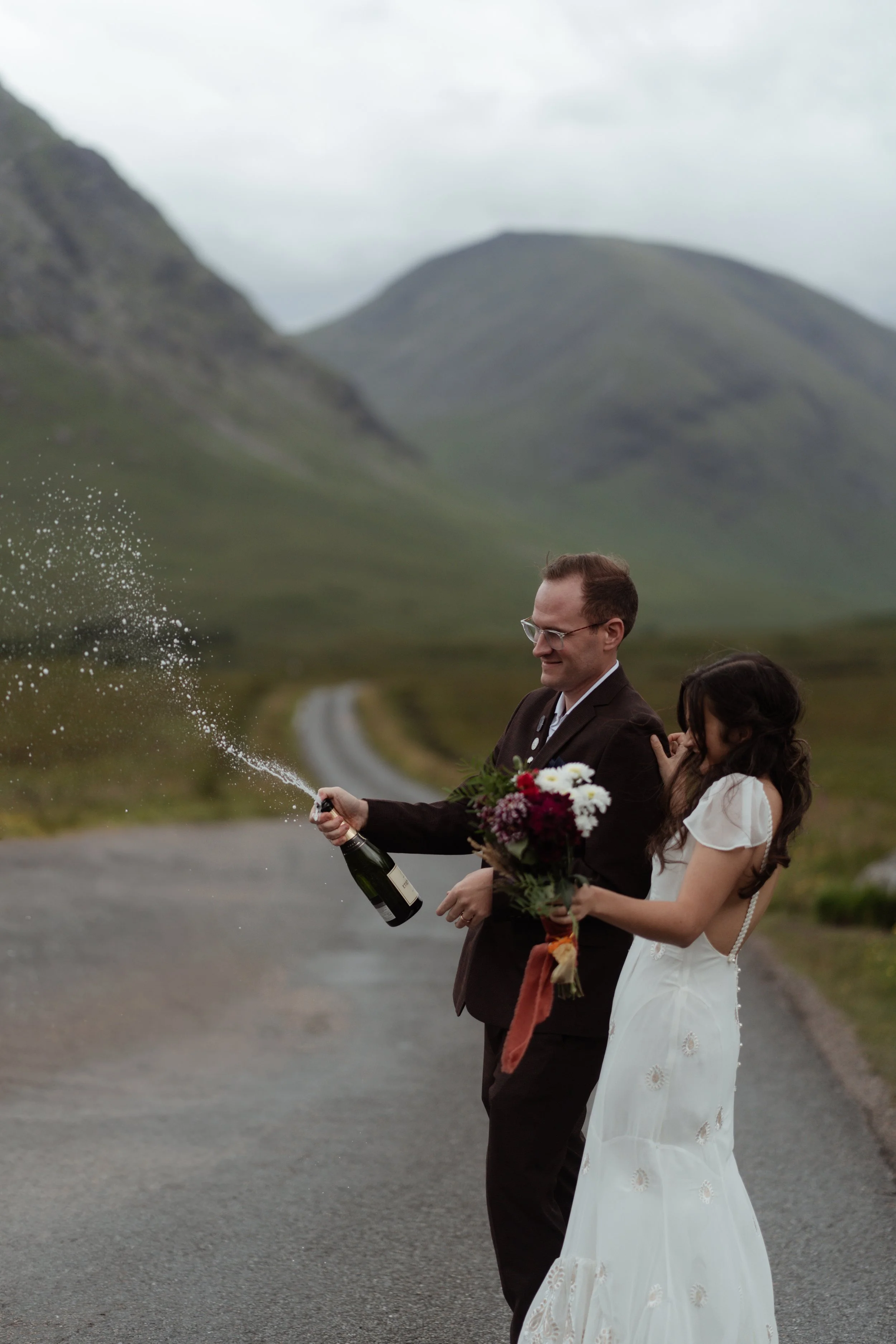 A couple dressed in wedding attire celebrating outdoors, with the man opening a champagne bottle and spraying it, while the woman holds a bouquet of flowers against a backdrop of green mountains and cloudy sky.