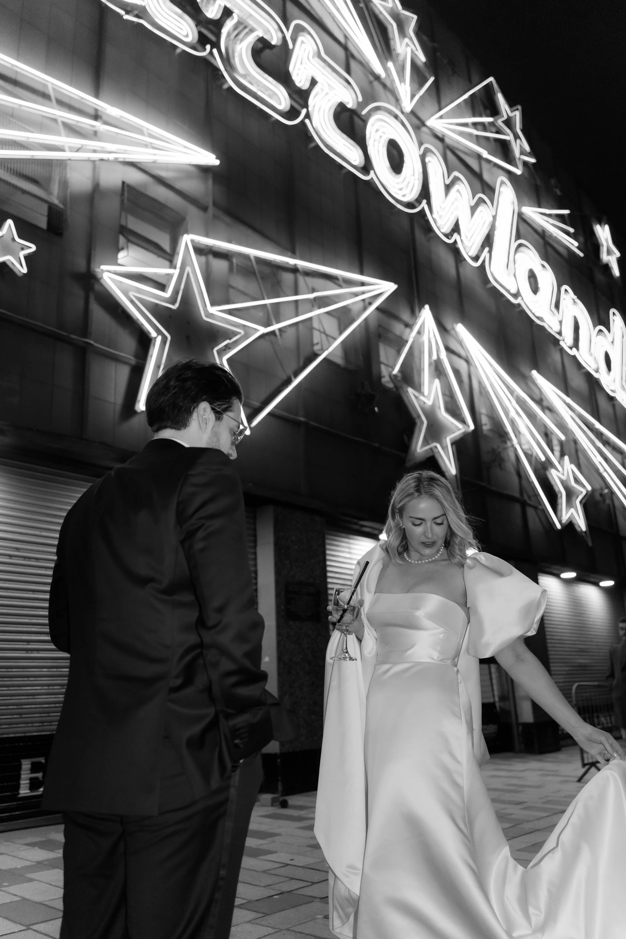 A black-and-white photo of a man in a suit and a woman in a wedding dress standing in front of a neon sign that reads 'Barrowlands' with star and shooting star decorations.