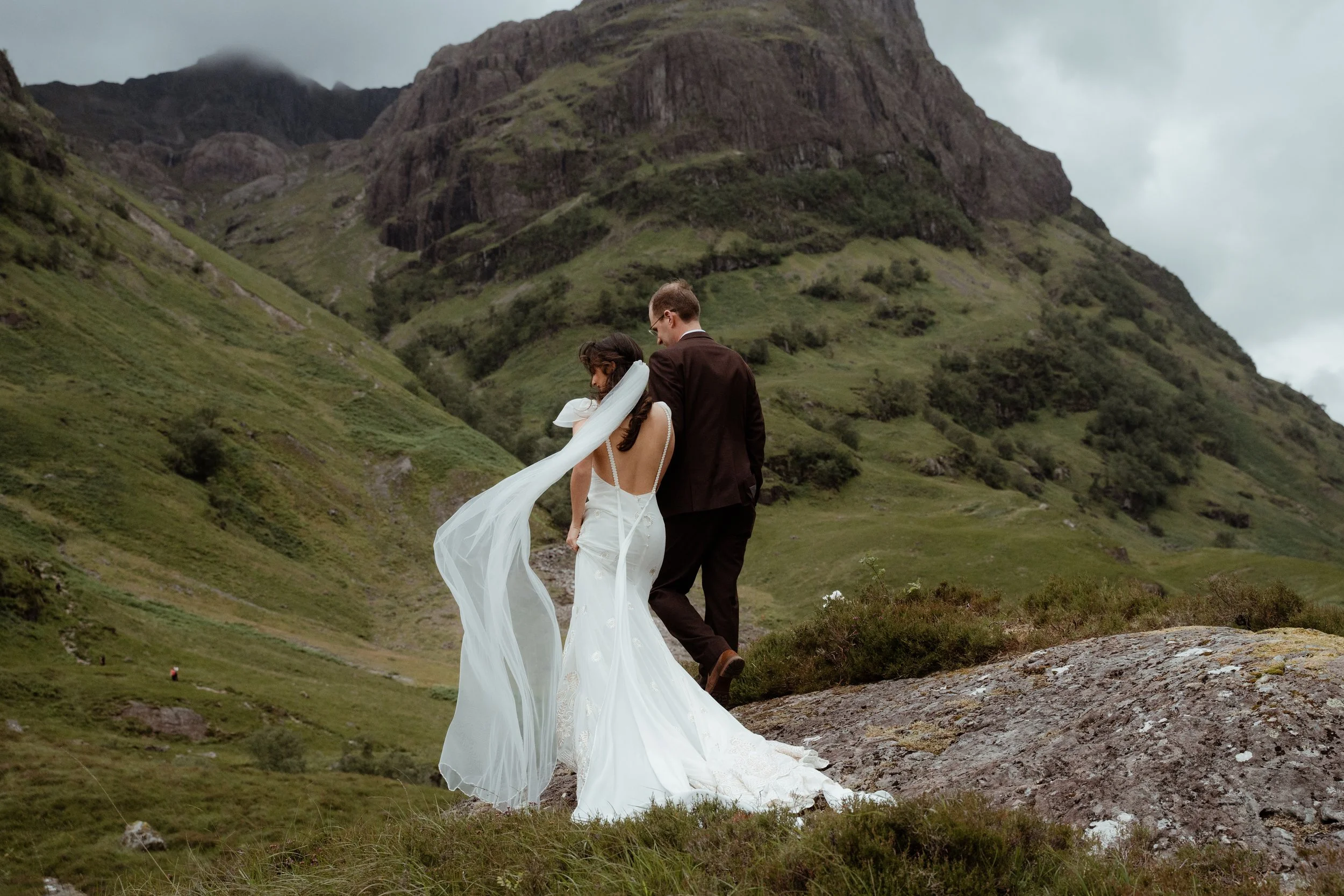 A bride and groom walking together on a rocky hillside with lush green mountains in the background.