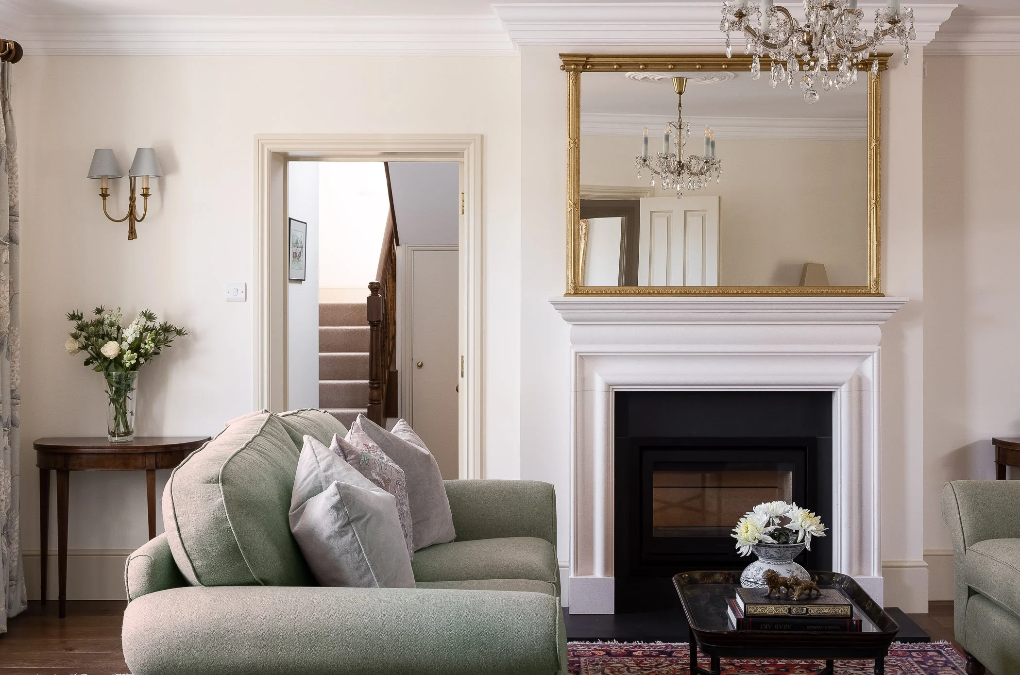 Living room with a pale green sofa, decorative pillows, a coffee table with flowers and books, a fireplace, a large mirror, crystal chandeliers, a wooden side table with a vase of flowers, a wall sconce, and an open doorway leading to a staircase.