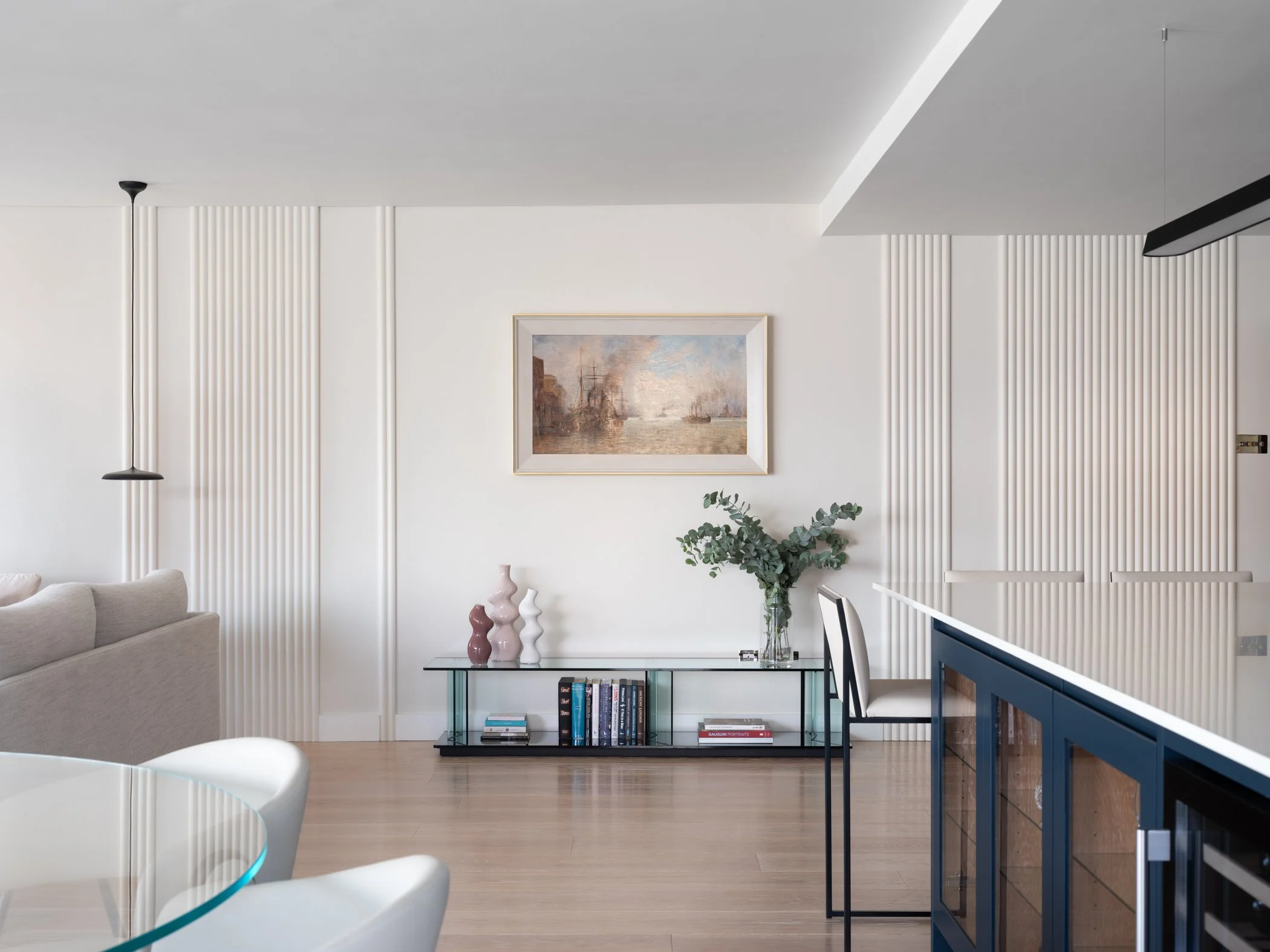 Modern living room with white walls, wooden flooring, a beige sofa on the left, a glass table with white chairs, a gallery wall with framed artwork, a black console table with vases and books, and a white bar counter with blue cabinets.