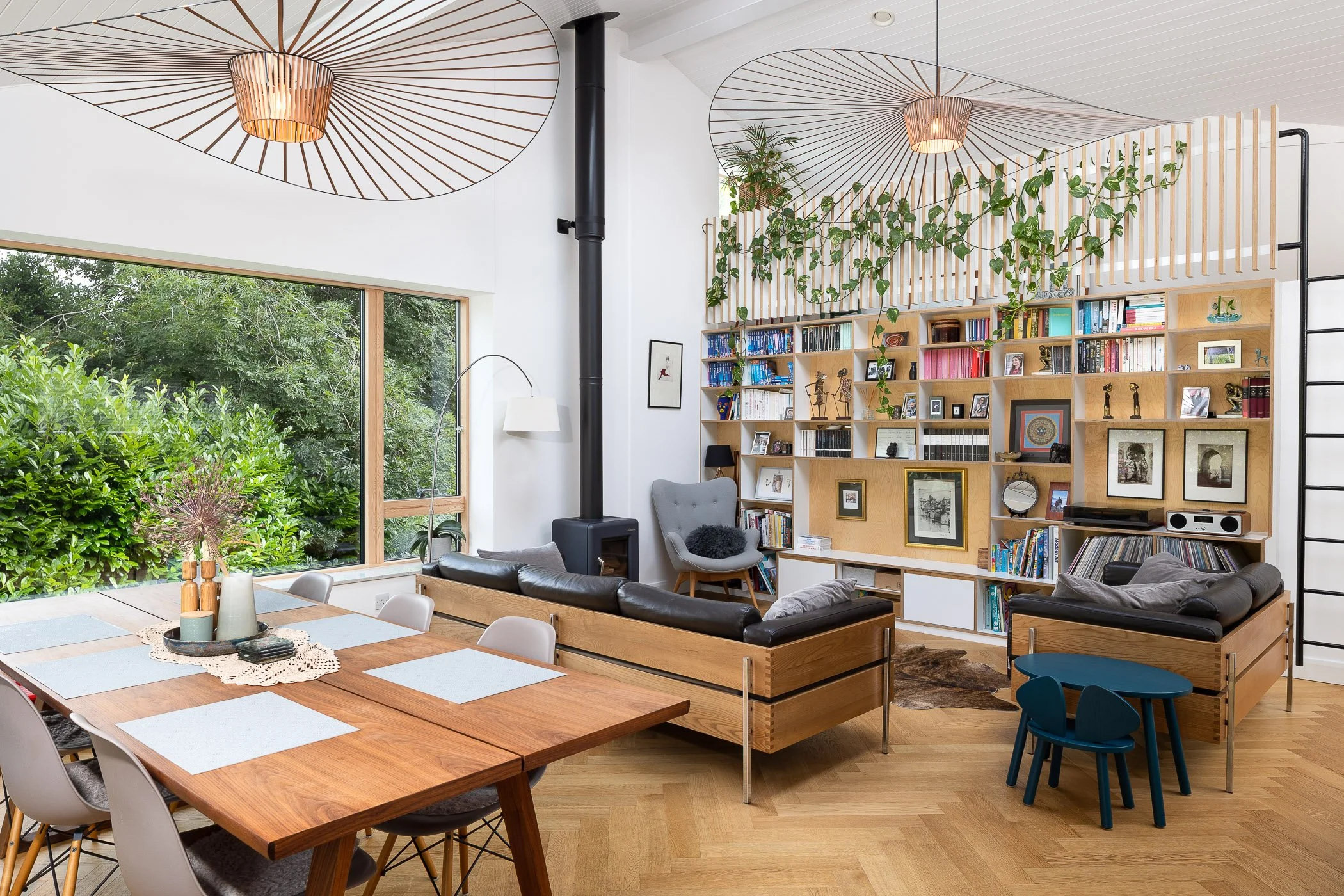 Modern living room with large window, wooden dining table with placemats, black leather sofas, a bookshelf with decorative items, and greenery on the ceiling and outside window.