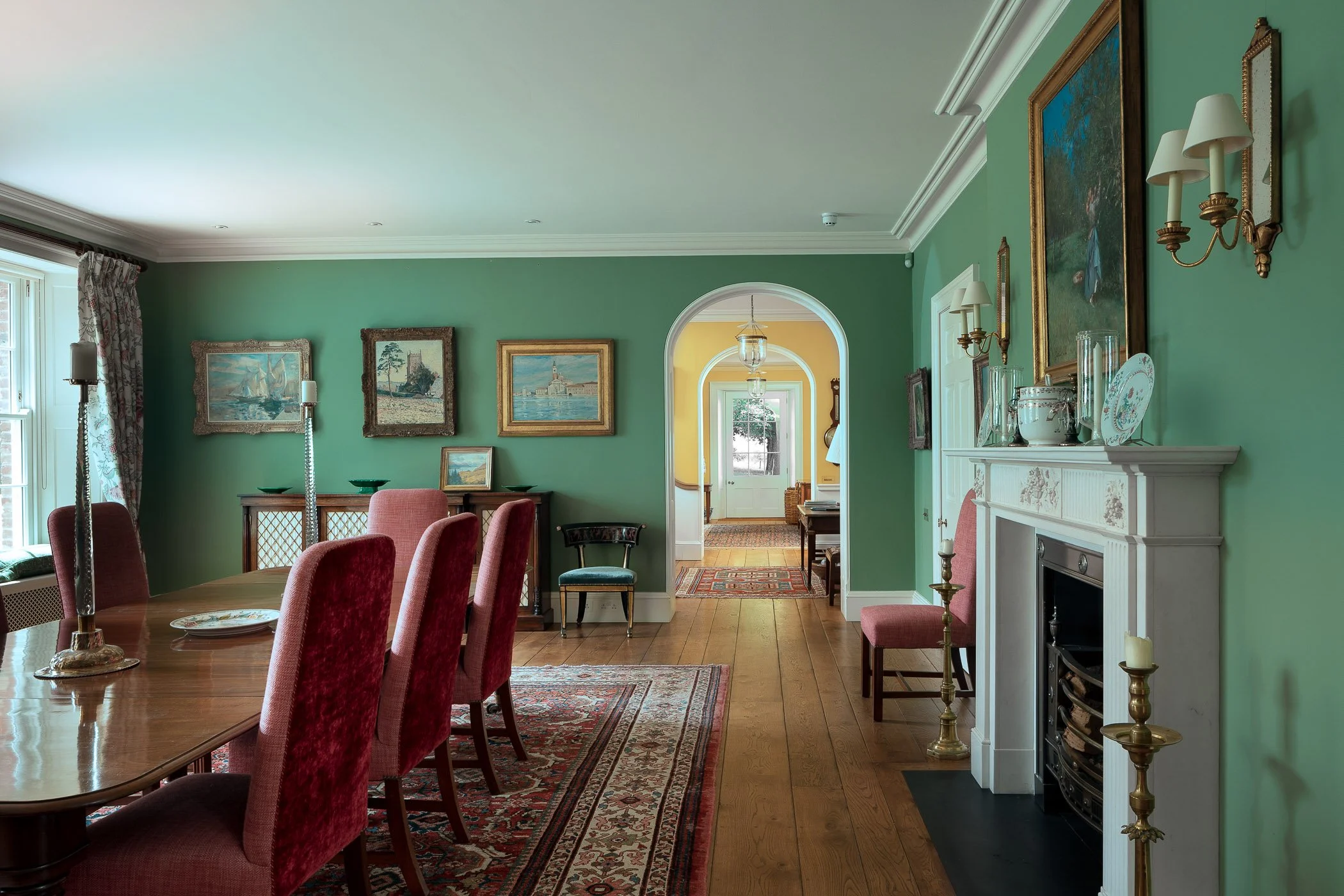 A dining room with green walls, a wooden table with red upholstered chairs, a fireplace, and artwork on the walls leading to a bright entryway.