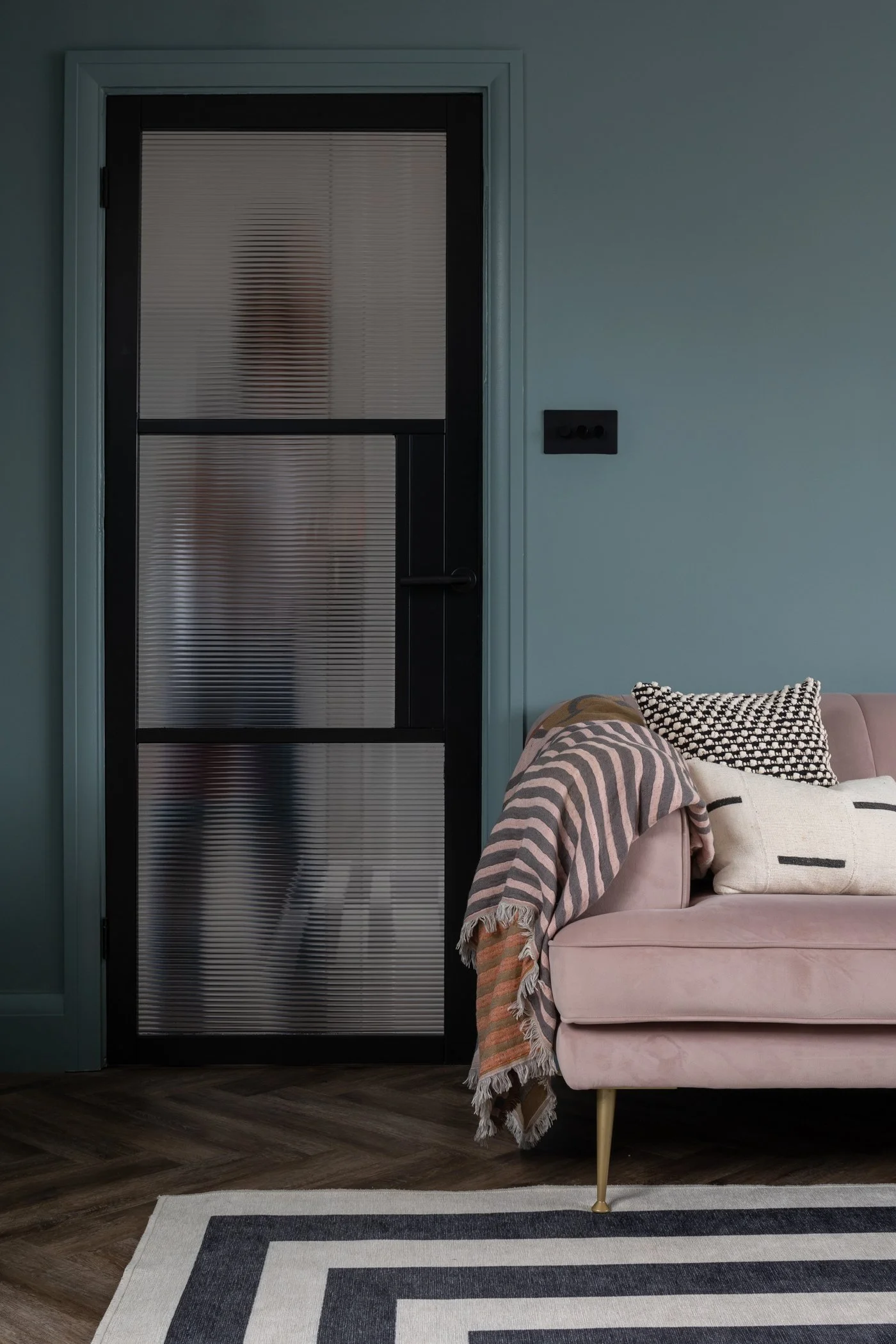 Room with a black glass door, pink sofa with striped and checked throw pillows, and a pink blanket on the armrest, against a teal wall, with a patterned rug and dark wood flooring.