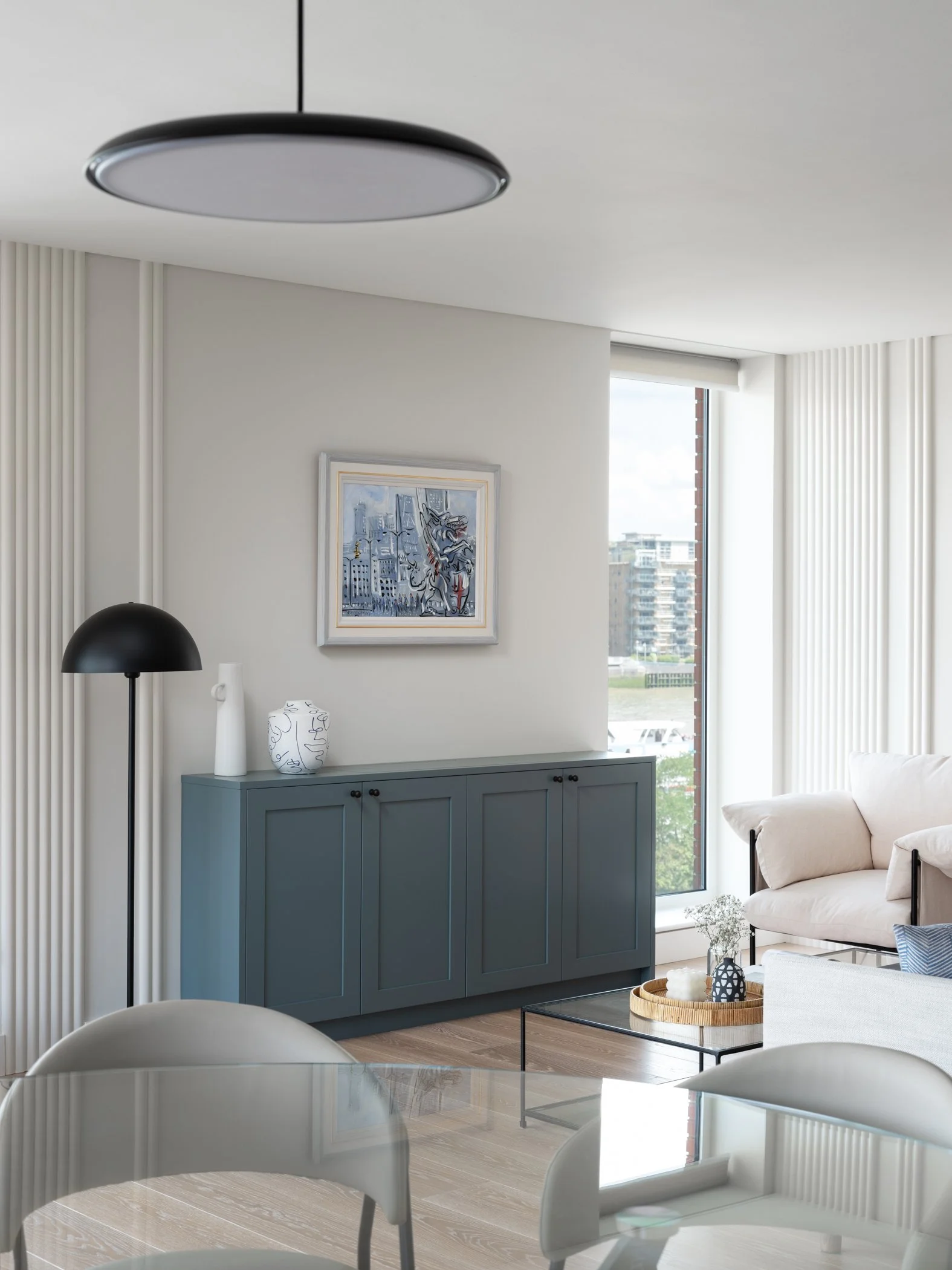 Modern living room with a white couch, blue sideboard, black floor lamp, glass coffee table, and wall art, featuring large windows and light-colored wood flooring.