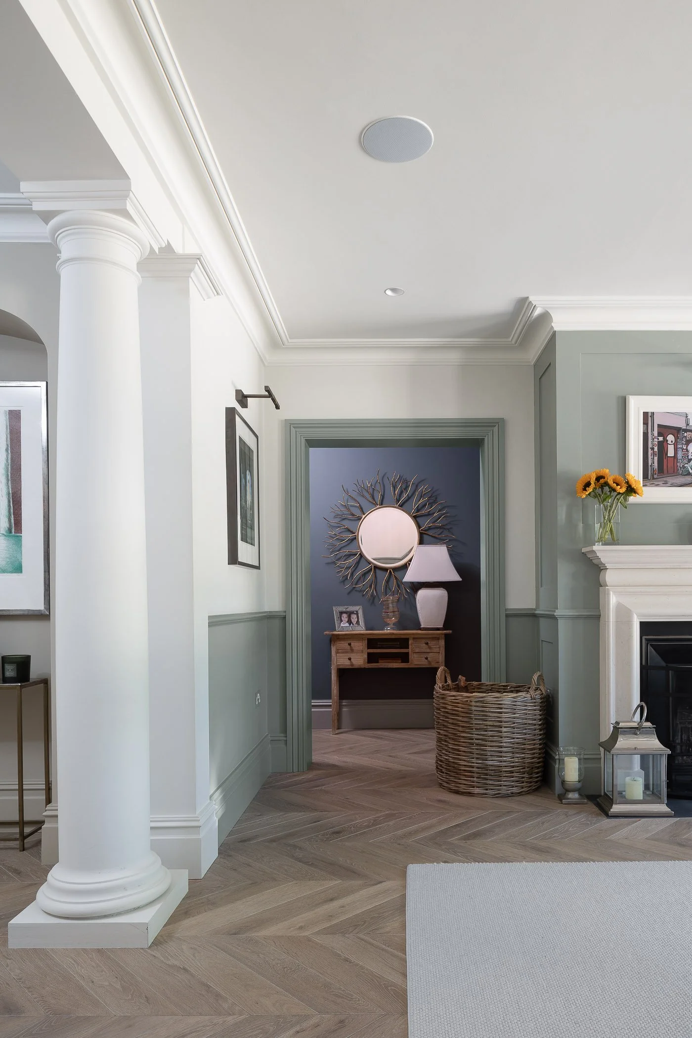 Living room with white and green walls, a white column, hardwood floor, and a doorway leading to a hallway with dark blue walls, a round mirror, a wooden console table, a lamp, and decorative items.