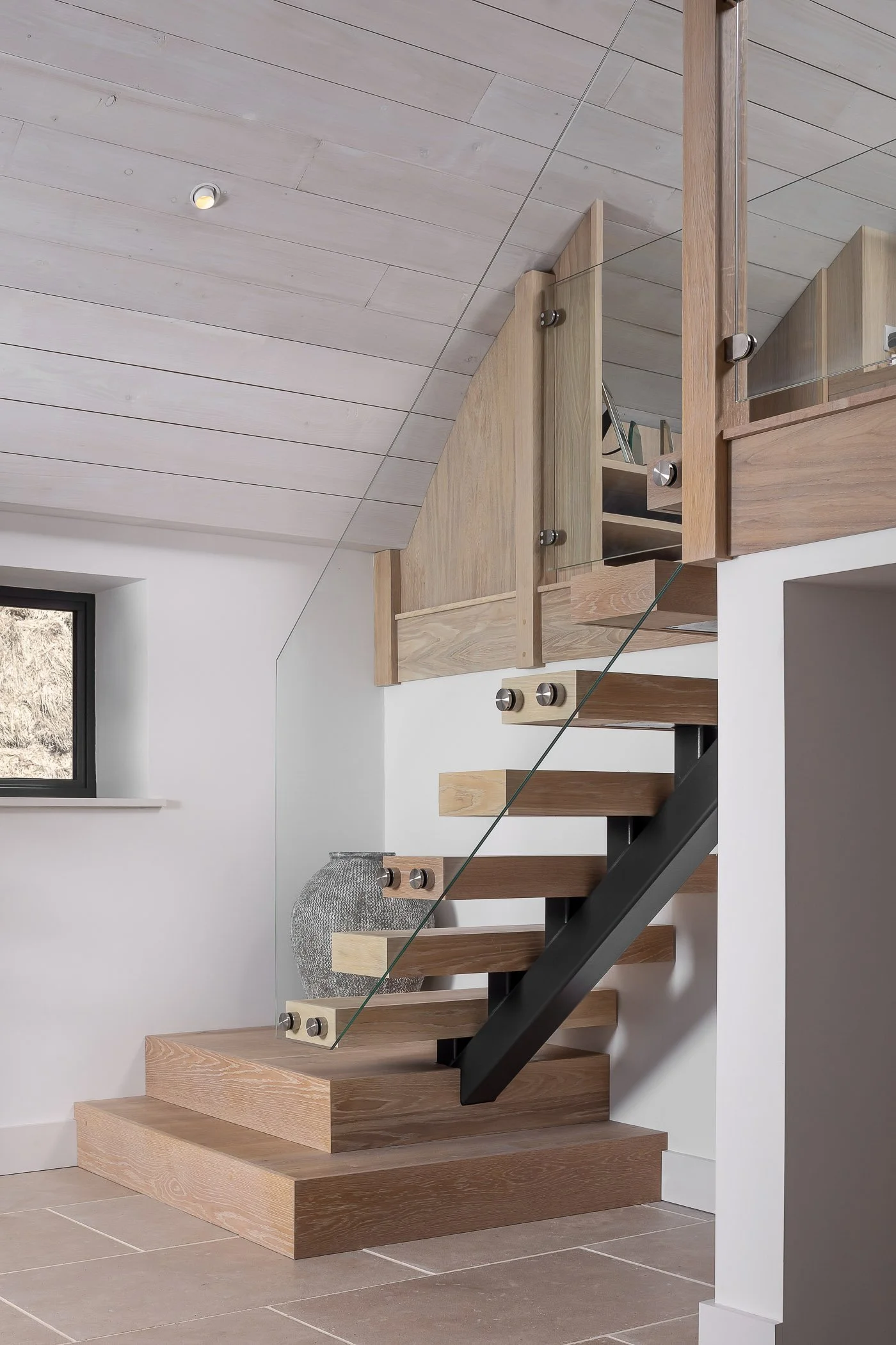 Interior view of a modern wooden staircase with glass railing and black support, leading to a mezzanine area with wooden railing, next to a small window in a room with white walls and light wood ceiling.