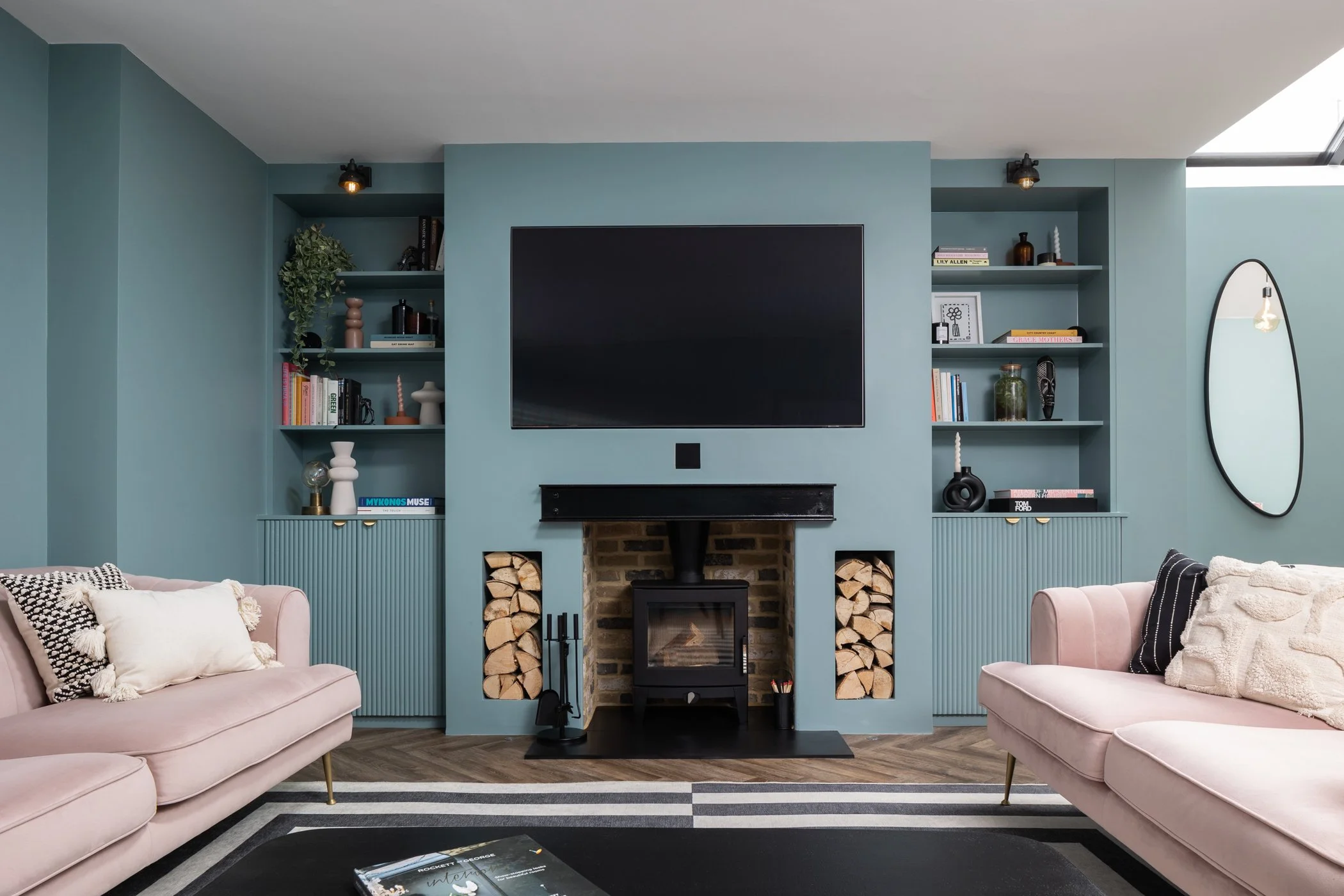 Living room with a teal wall, a mounted TV above a wood-burning stove with firewood on both sides, two pastel pink sofas with black and white pillows, built-in teal bookshelves with books and decor, a wall mirror, and a skylight.
