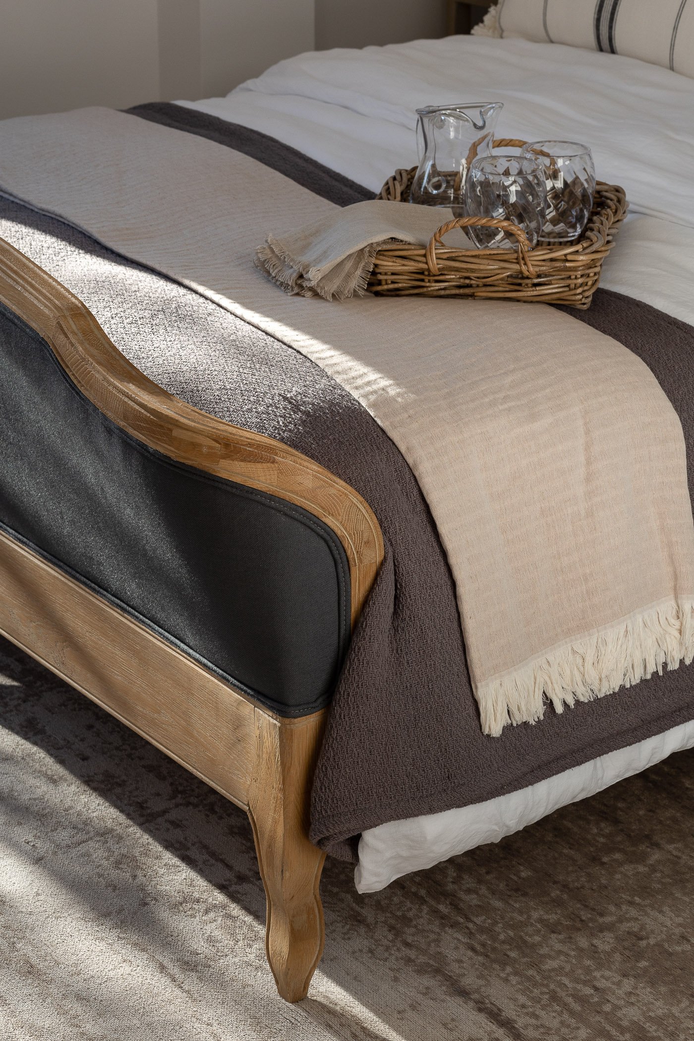 A neatly made bed with a beige and brown bedspread, topped with a wicker tray holding glassware and a pitcher, and a folded beige cloth on the tray, in a cozy bedroom setting.