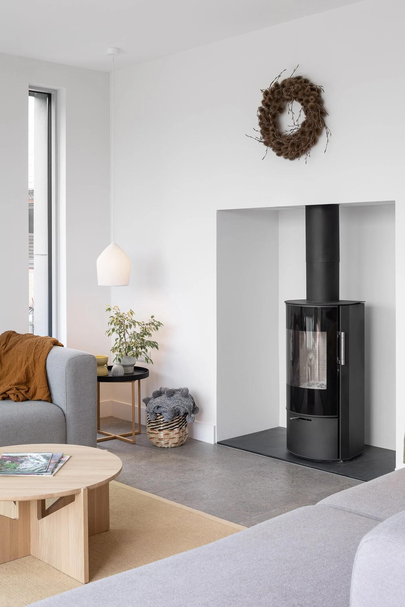 Living room with a wood stove, gray sofas, a round wooden coffee table, a side table with a potted plant, and a decorative wreath above the fireplace.