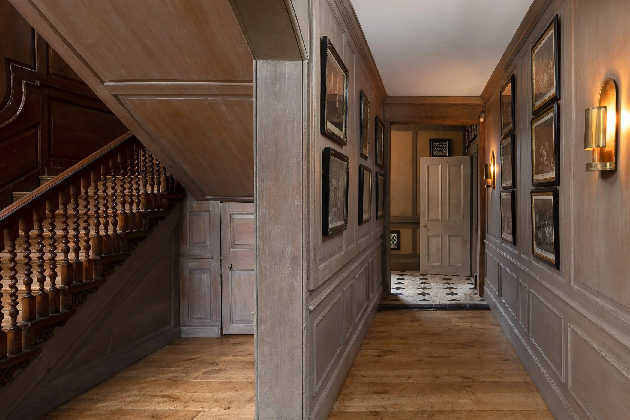 Interior view of a wooden hallway with framed artwork, wall-mounted sconces, and a staircase on the left.