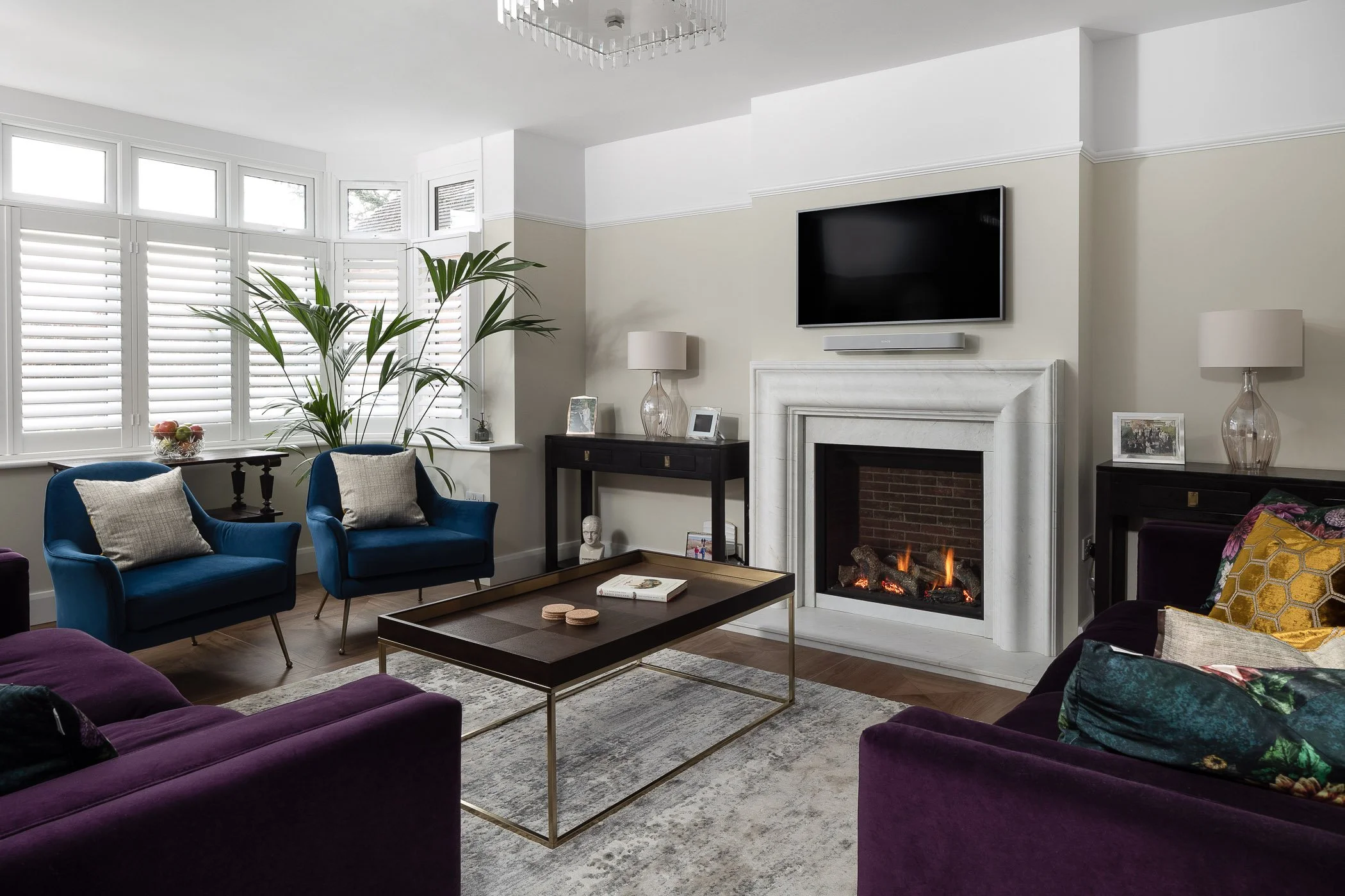 Living room with fireplace, flat-screen TV, two blue armchairs, purple sofa, coffee table, lamps, and large window with white blinds.