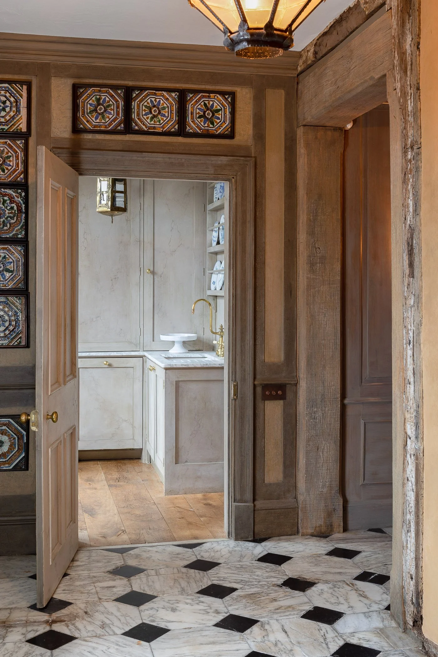 View of a doorway leading into a small bathroom with a white sink and a gold faucet, partially visible behind a door, with black and white patterned marble flooring and wooden trim.