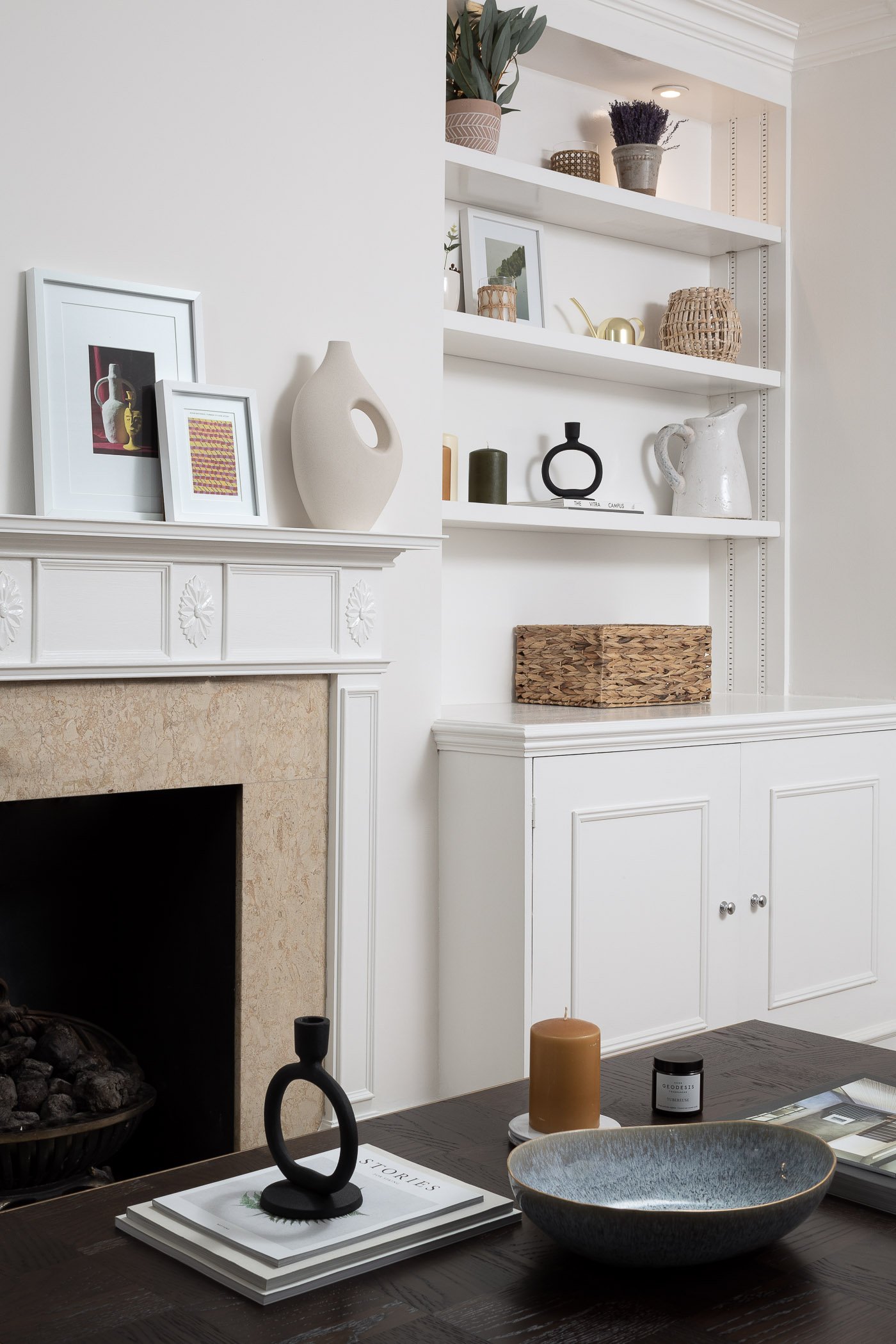 Interior of a living room with white built-in shelves displaying decorative items, a fireplace with a beige stone surround, and a dark wooden table with candles, a bowl, and magazines.