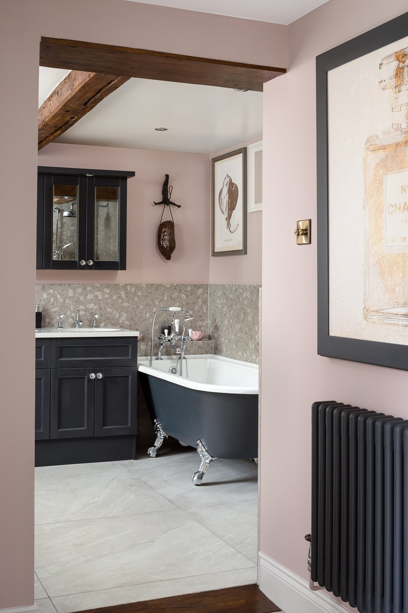 Interior view of a bathroom with a vintage clawfoot bathtub, dark gray cabinet, framed artwork, and decorative wall hanging, with light pink walls, a wooden beam ceiling detail, and beige tiled flooring.