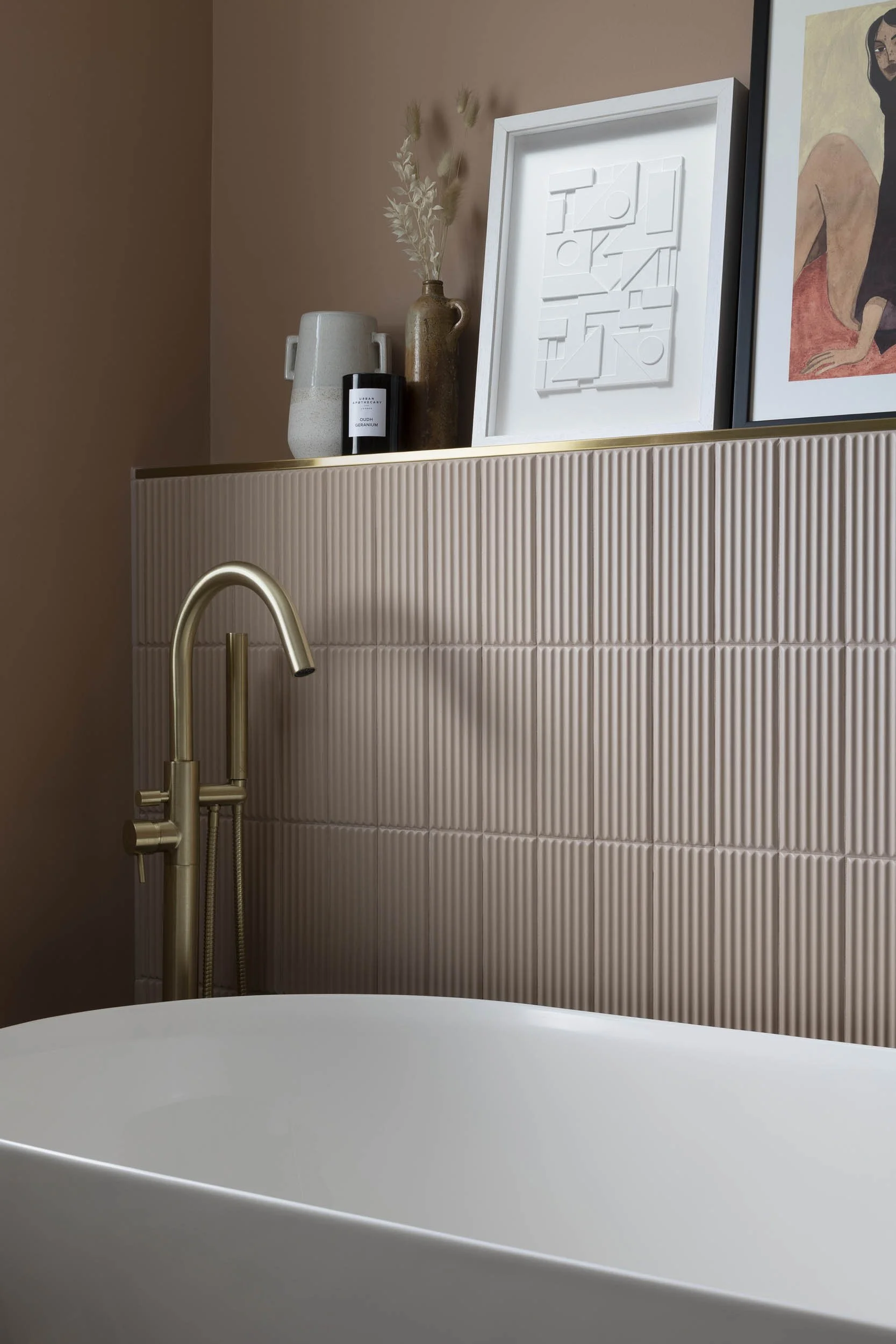Close-up of a modern bathtub with a gold faucet, a textured beige tiled wall, and decorative items including vases and framed artwork on a shelf above the tub.
