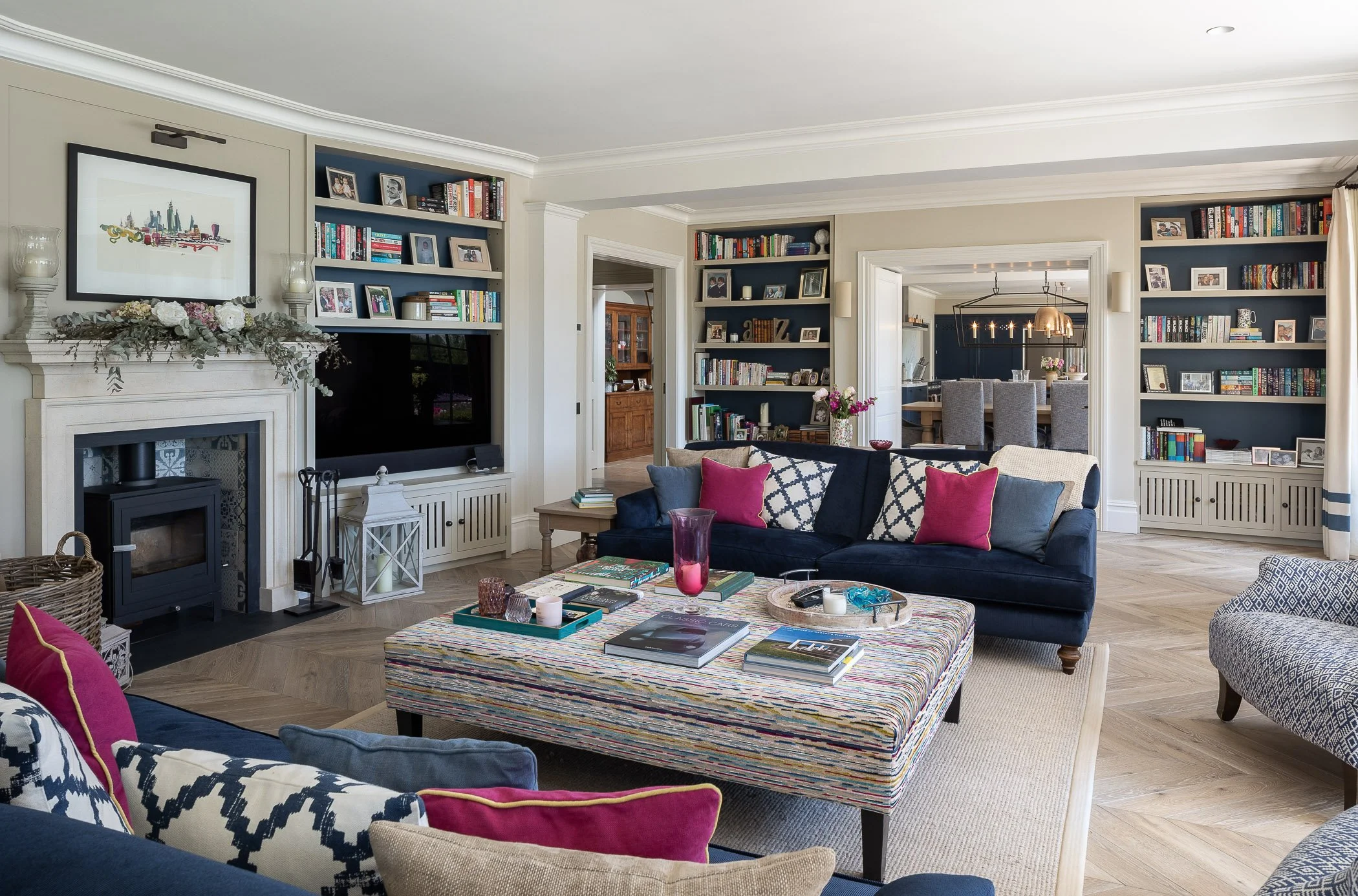 Living room with a fireplace, bookshelves, a dark blue sofa with pink and patterned cushions, a colorful ottoman with books and candles, and a wall-mounted TV.