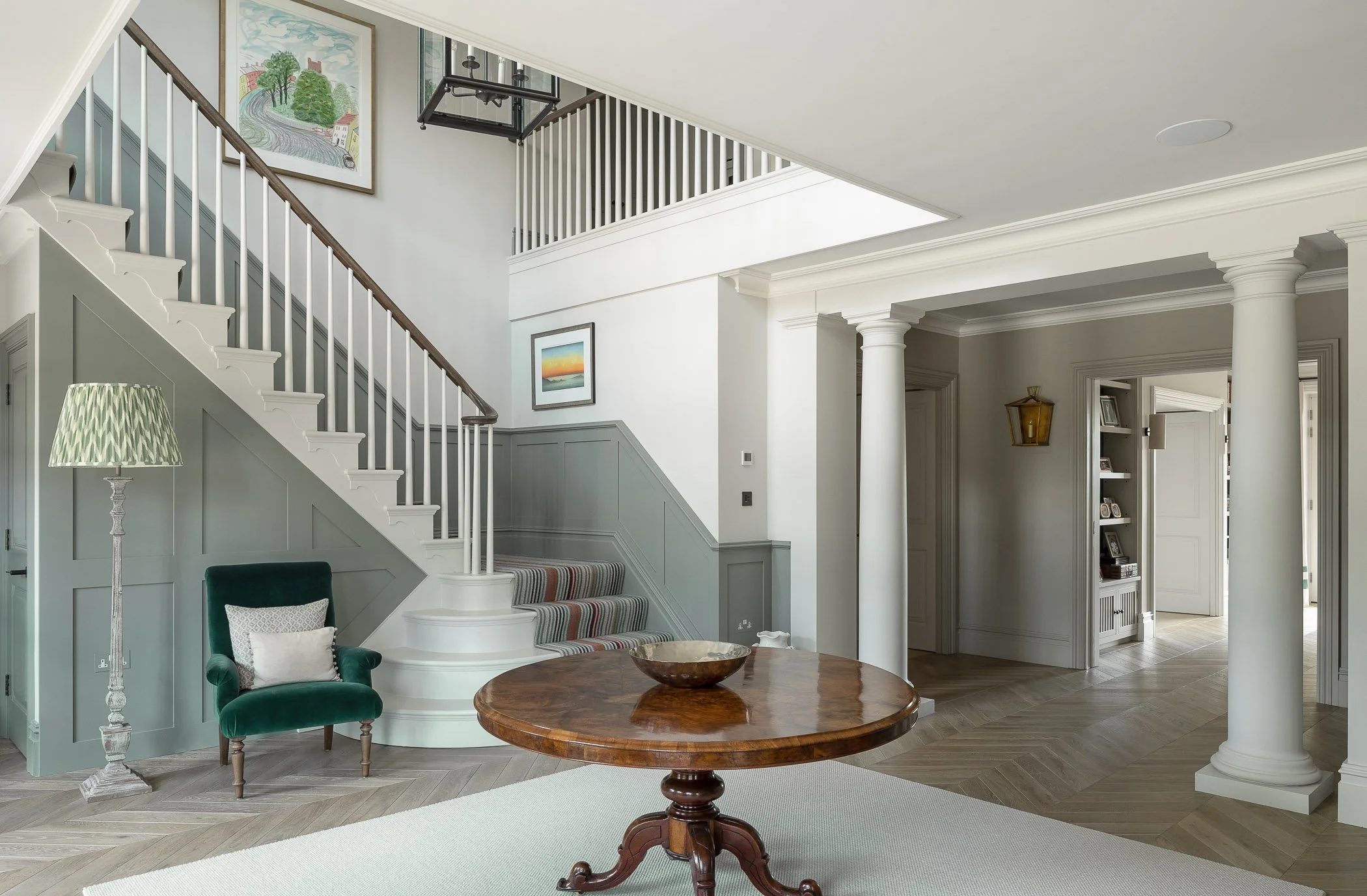 Interior view of a spacious, elegantly decorated living area with a staircase, armchair, round wooden table, and decorative columns.