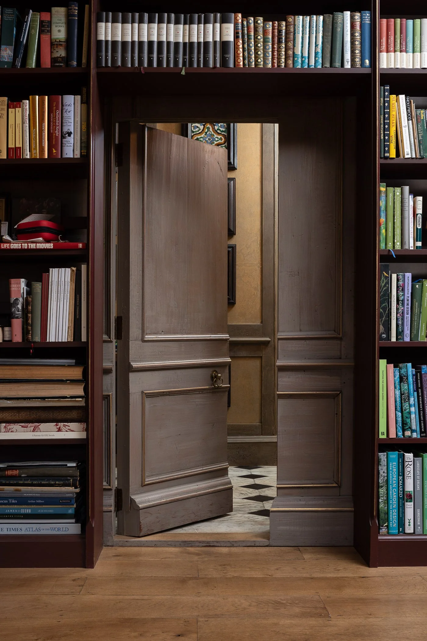 View through an open wooden door in a library surrounded by bookshelves filled with books. The floor is wooden, and part of a patterned tiled floor is visible through the doorway.