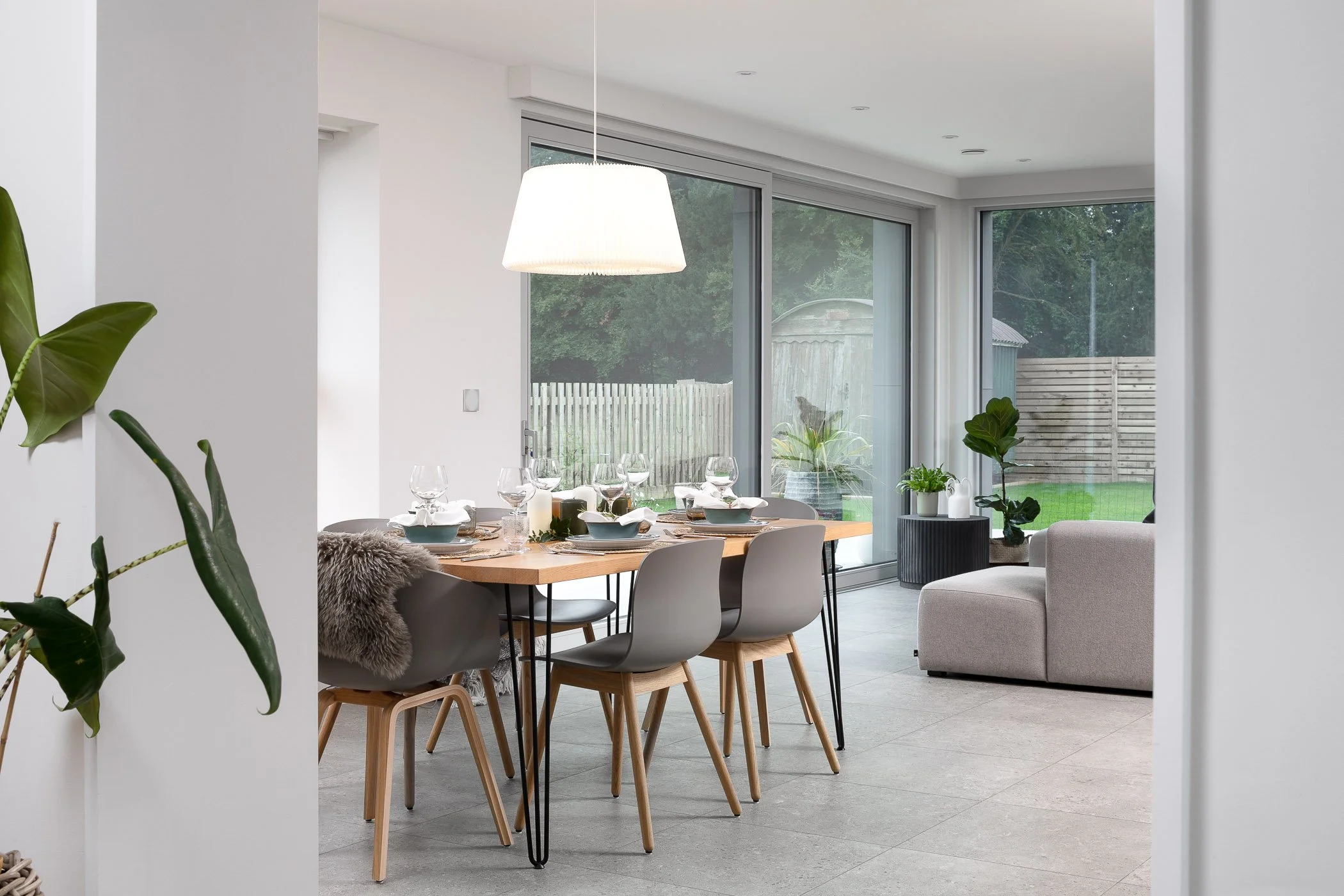 Modern dining area with a wooden table set for a meal, surrounded by light gray chairs, with sliding glass doors leading to a backyard with trees, lawn, and a wooden fence, decorated with potted plants and a gray armchair.