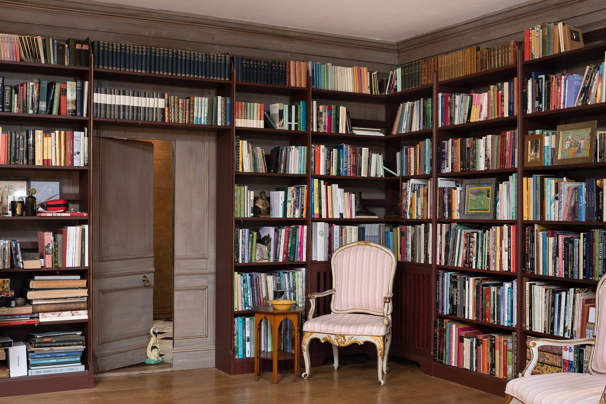 A cozy library corner with tall wooden bookshelves filled with books, a pink striped upholstered armchair, a small wooden side table with a bowl, and a doorway leading to another room.