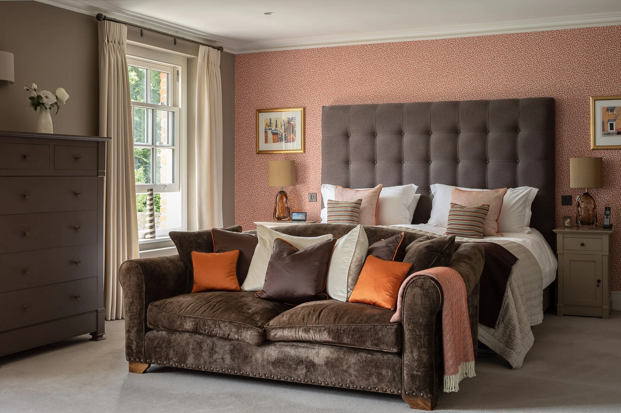 A bedroom with a bed, a sofa at the foot of the bed, a window with cream curtains, and a dresser. The room is decorated with framed artwork, lamps, and pillows in warm tones.