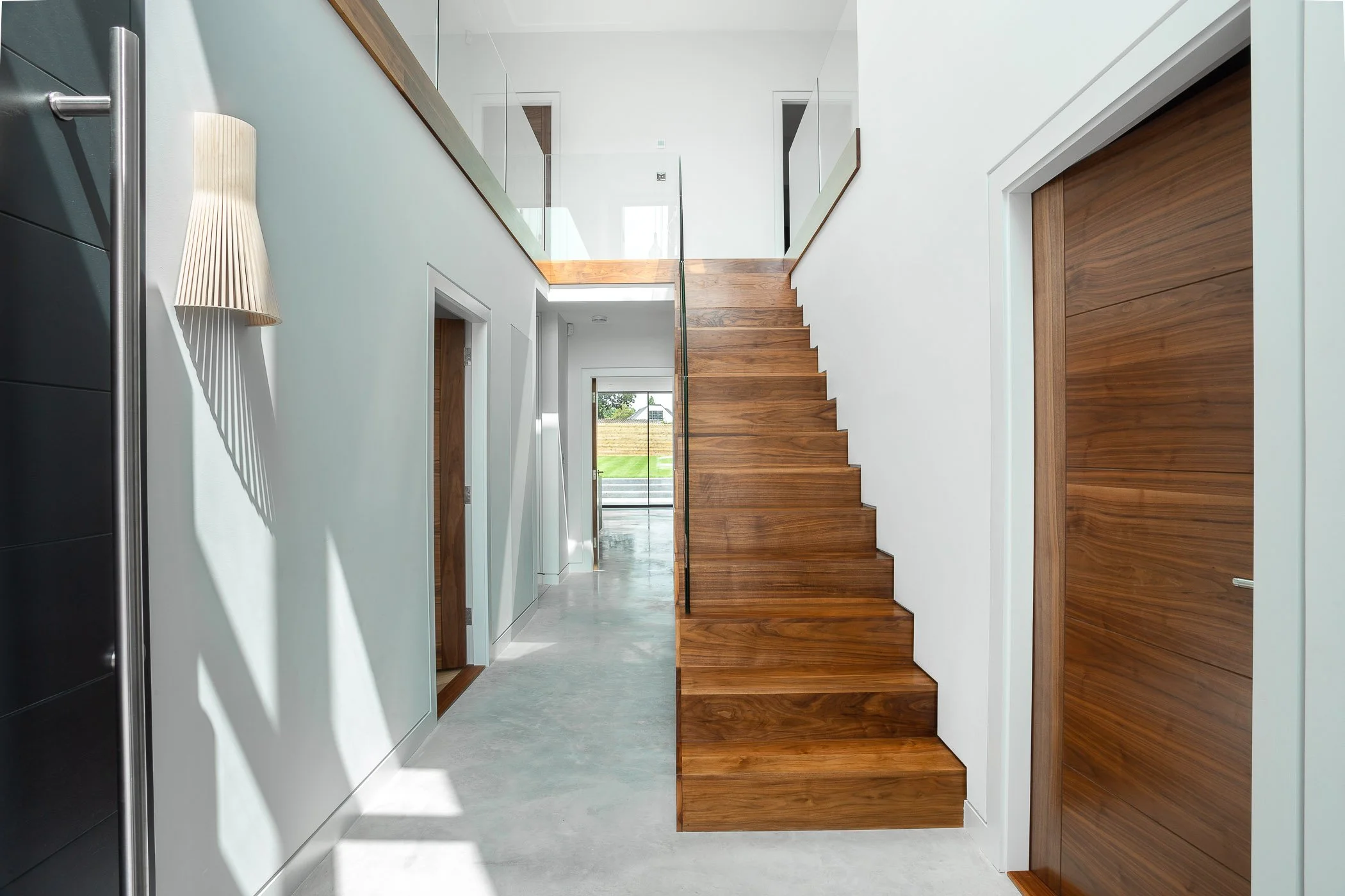 Modern interior of a house with white walls, wooden staircase, and glass railings leading to an upper floor with landscape visible through large windows at the end of the hallway.