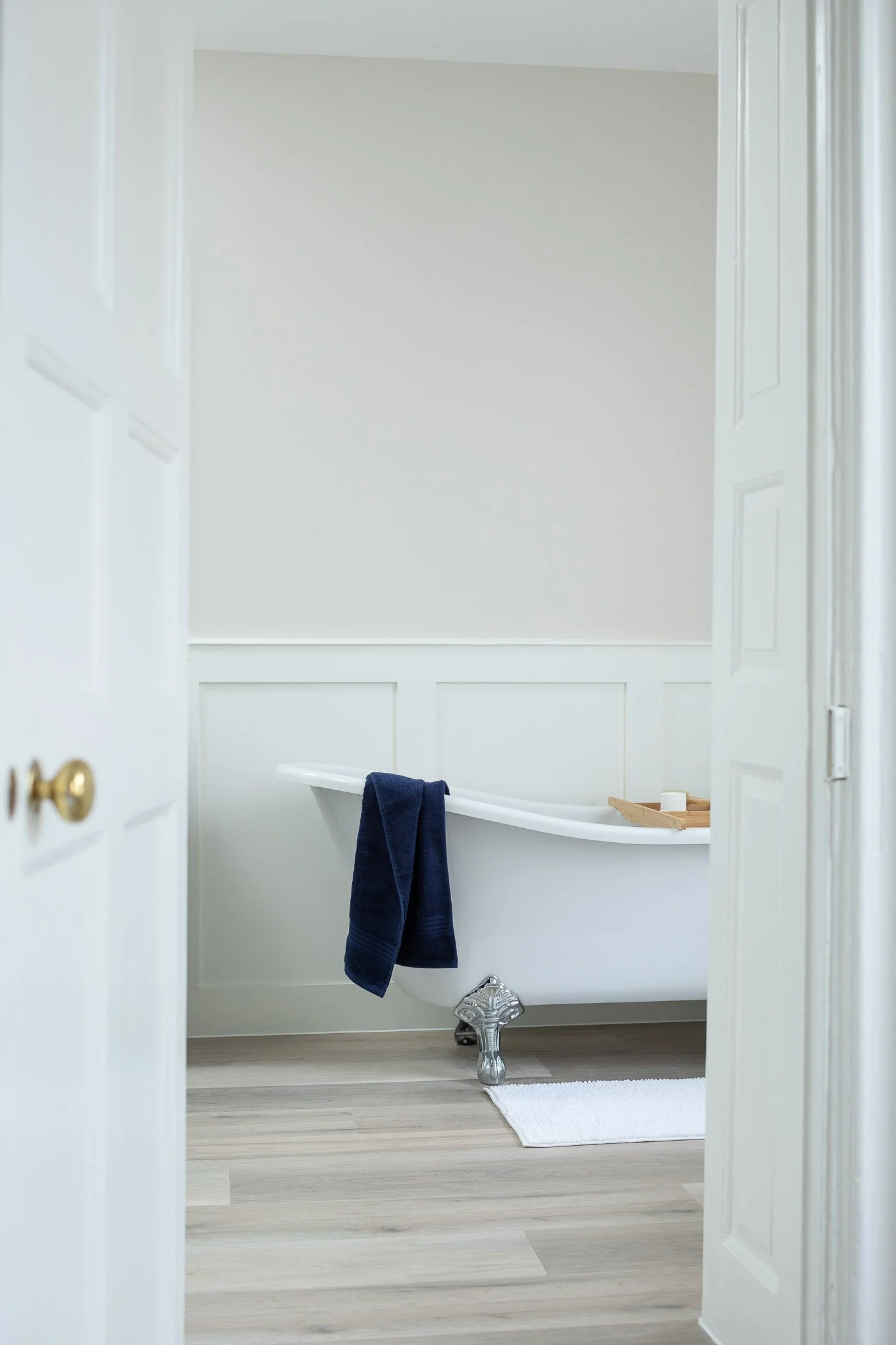Empty bathroom with a vintage clawfoot bathtub, navy towel hanging on the side, wooden tray with soap, and white rug on light wood floor, viewed through partially open white doors.