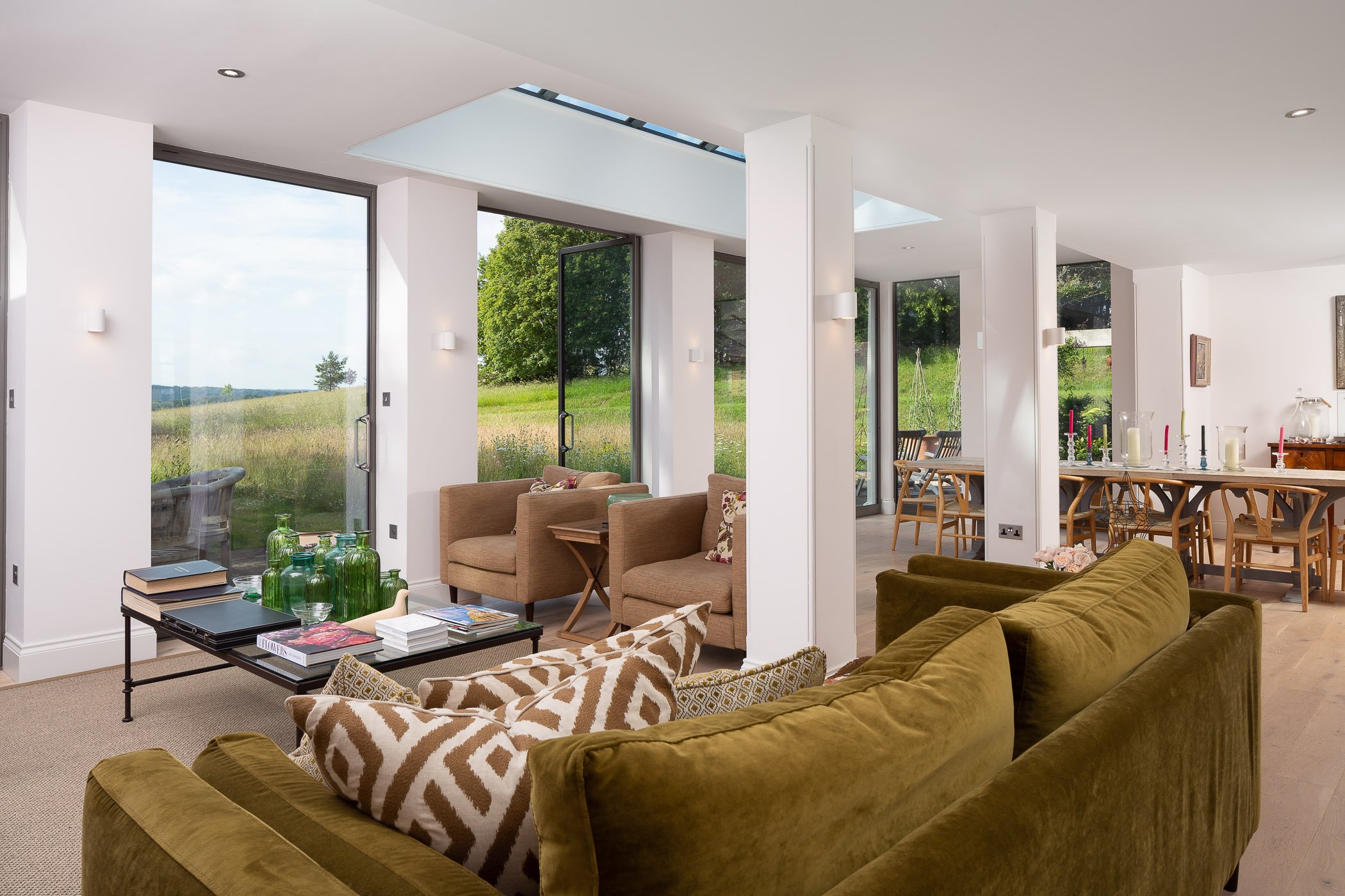 Living room with large windows and view of green outdoor landscape, beige and green furniture, and wooden dining table with chairs.