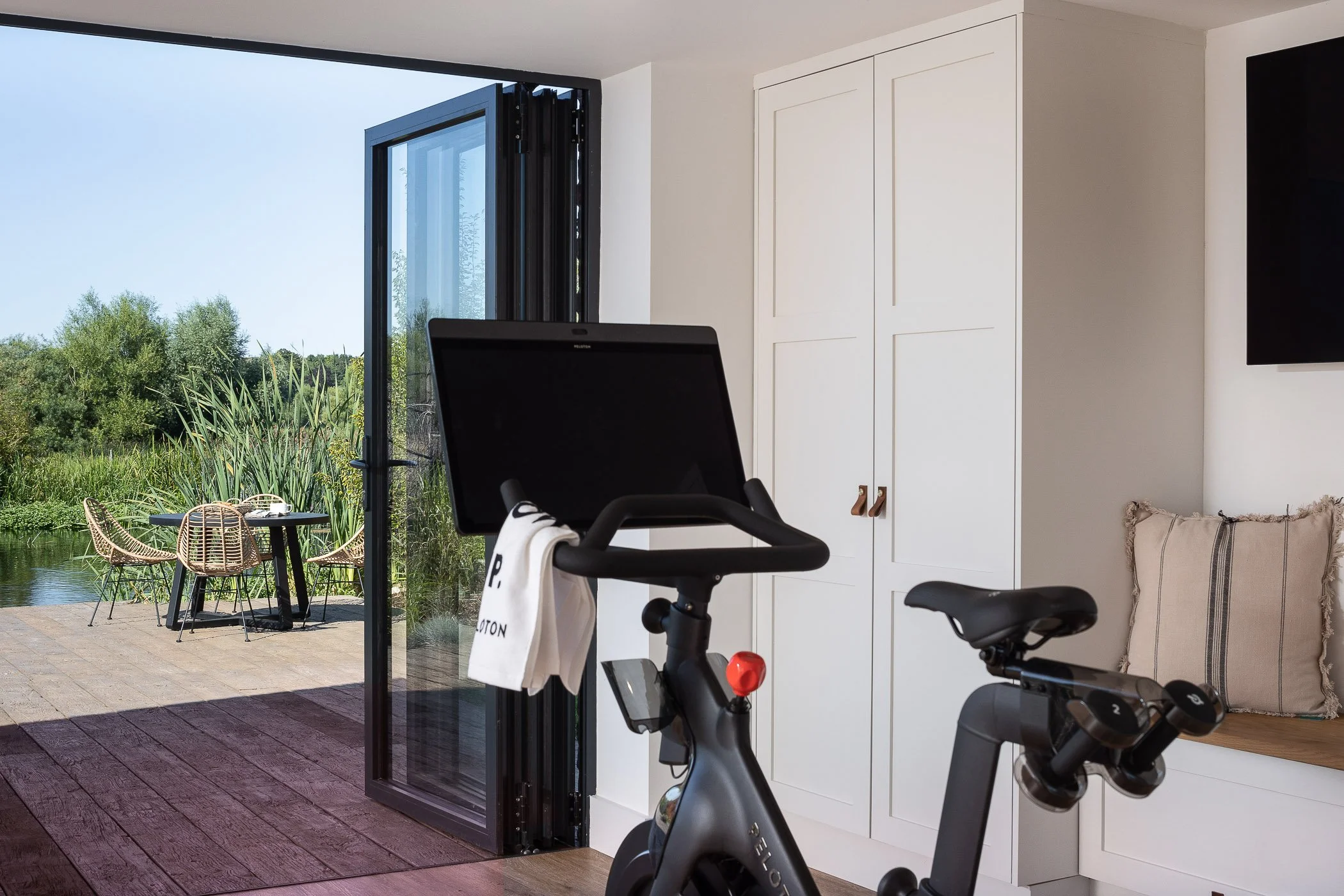 Indoor exercise bike facing an open balcony door with a view of an outdoor patio and green landscape with water and trees.