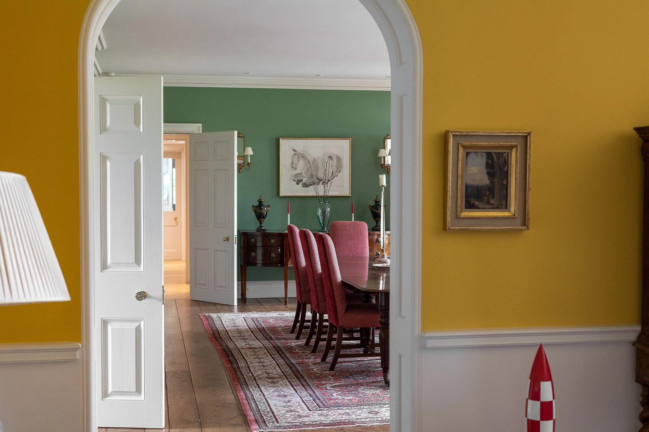 View through an arched doorway into a dining room with green walls, a wooden table with red upholstered chairs, a rug, and a sideboard with vases and framed artwork.