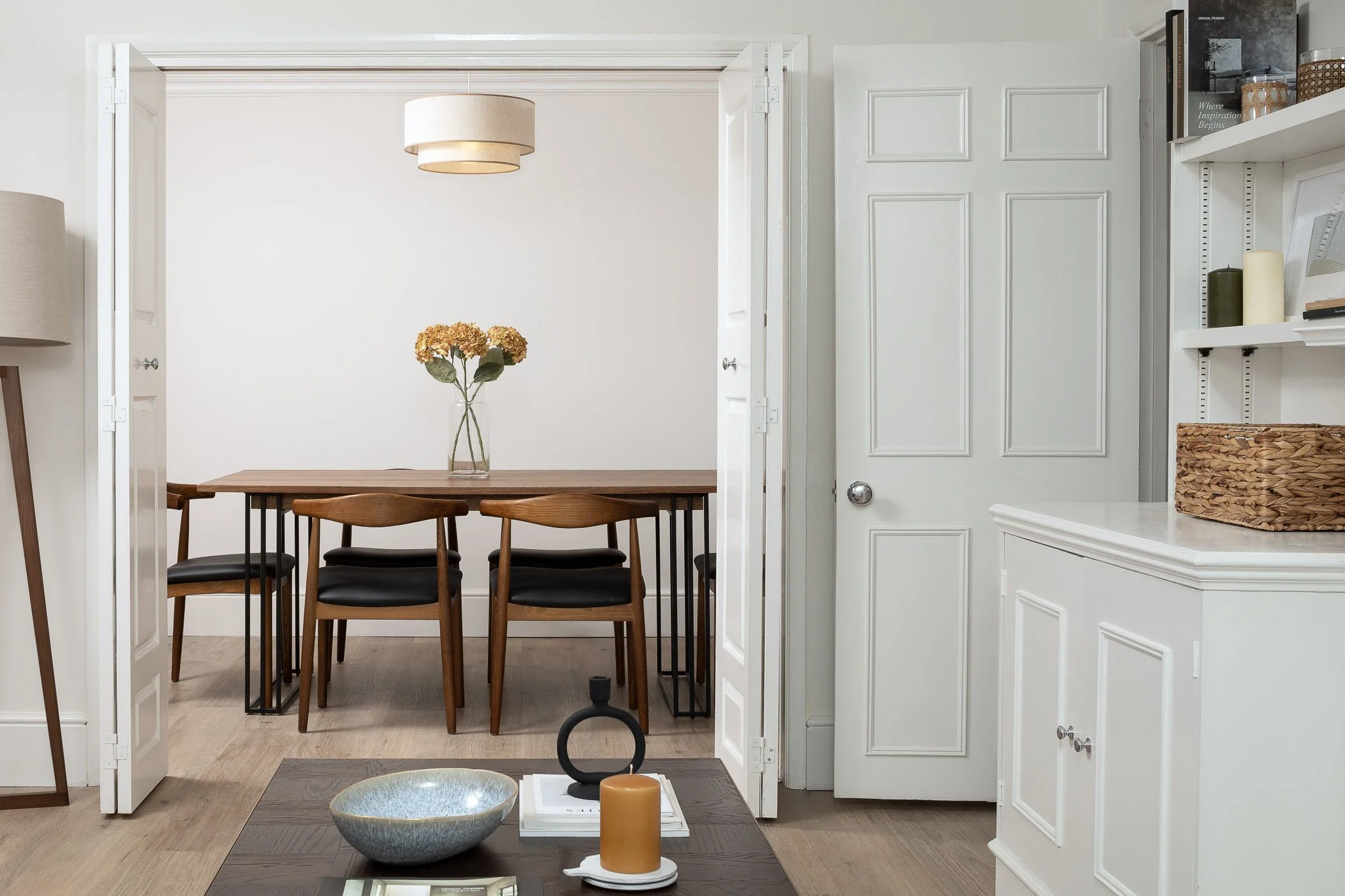 A dining room with a wooden table, four chairs, a vase with flowers, white walls, a hanging light, and a white door.