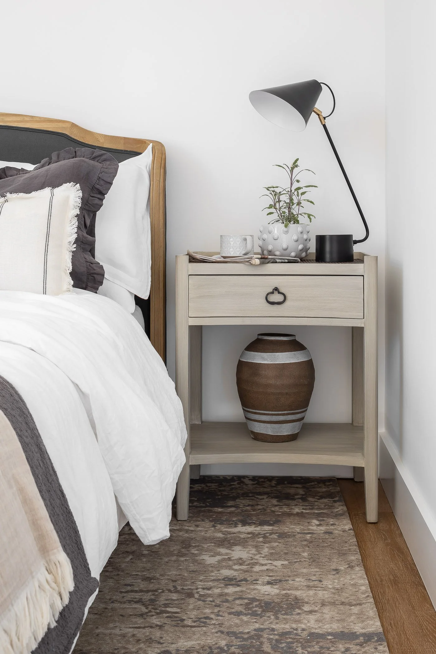 A bedroom nightstand next to a bed with white and dark gray bedding, a small potted plant, a white mug, some magazines, a black table lamp, and a large decorative brown and white vase underneath.