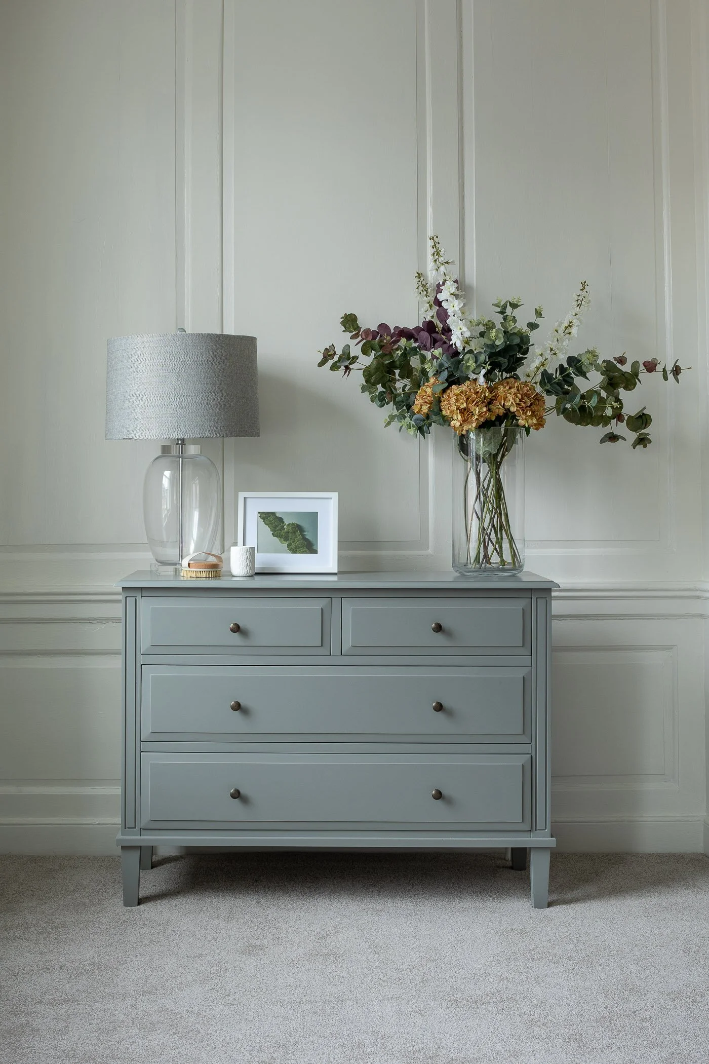 A gray dresser with five drawers, decorated with a glass lamp, a framed photo, a white cup, a small wooden tray, and a tall vase with flowers, against a white wall.