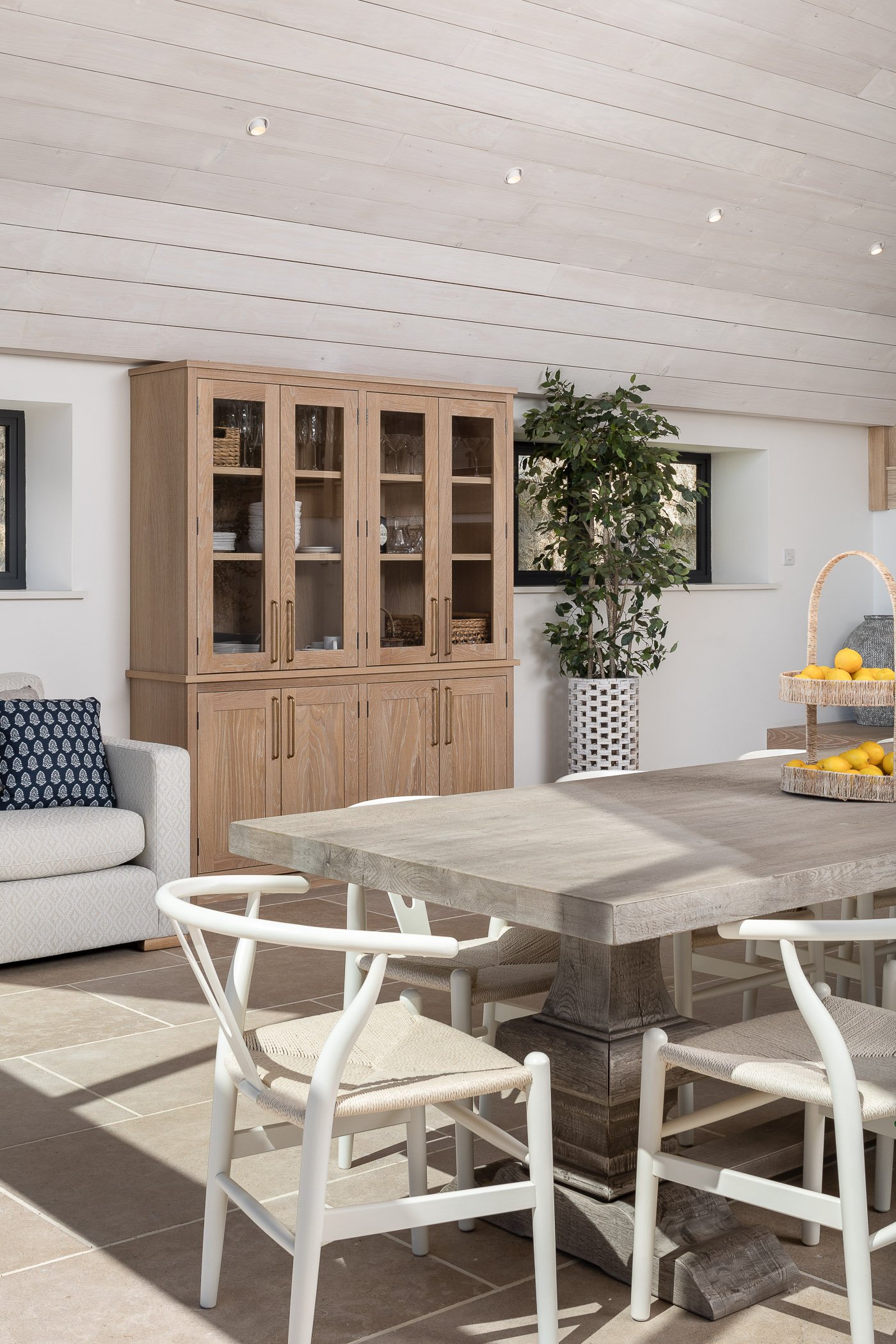 A dining area with a large wooden table and six white chairs, a wooden glass-fronted cabinet, a white sofa with a patterned pillow, a green potted plant, and a small window in a bright room with a sloped ceiling.