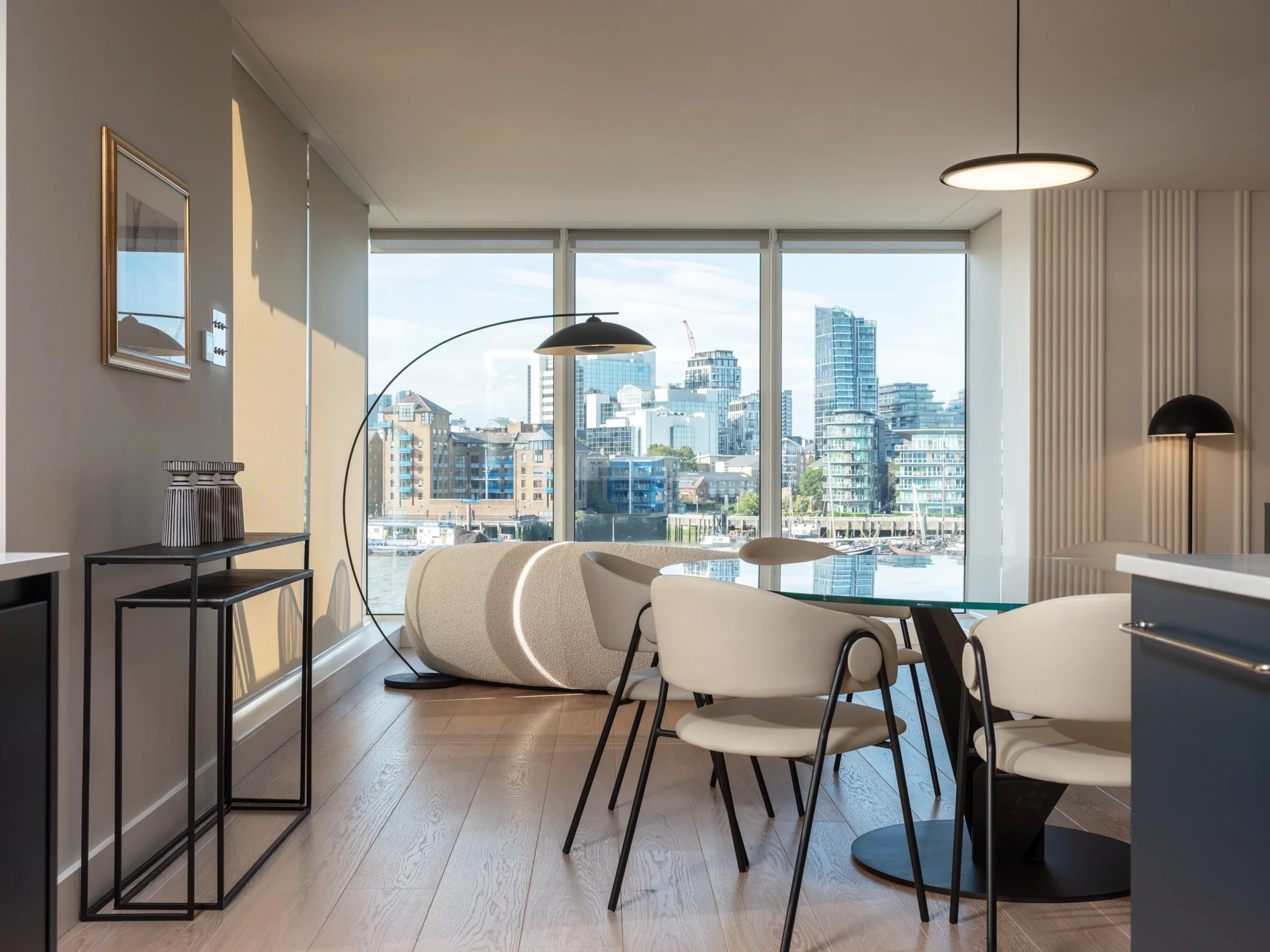 Modern living room with large windows overlooking a city skyline, white chairs around a glass dining table, black floor lamp, and minimalist decor.