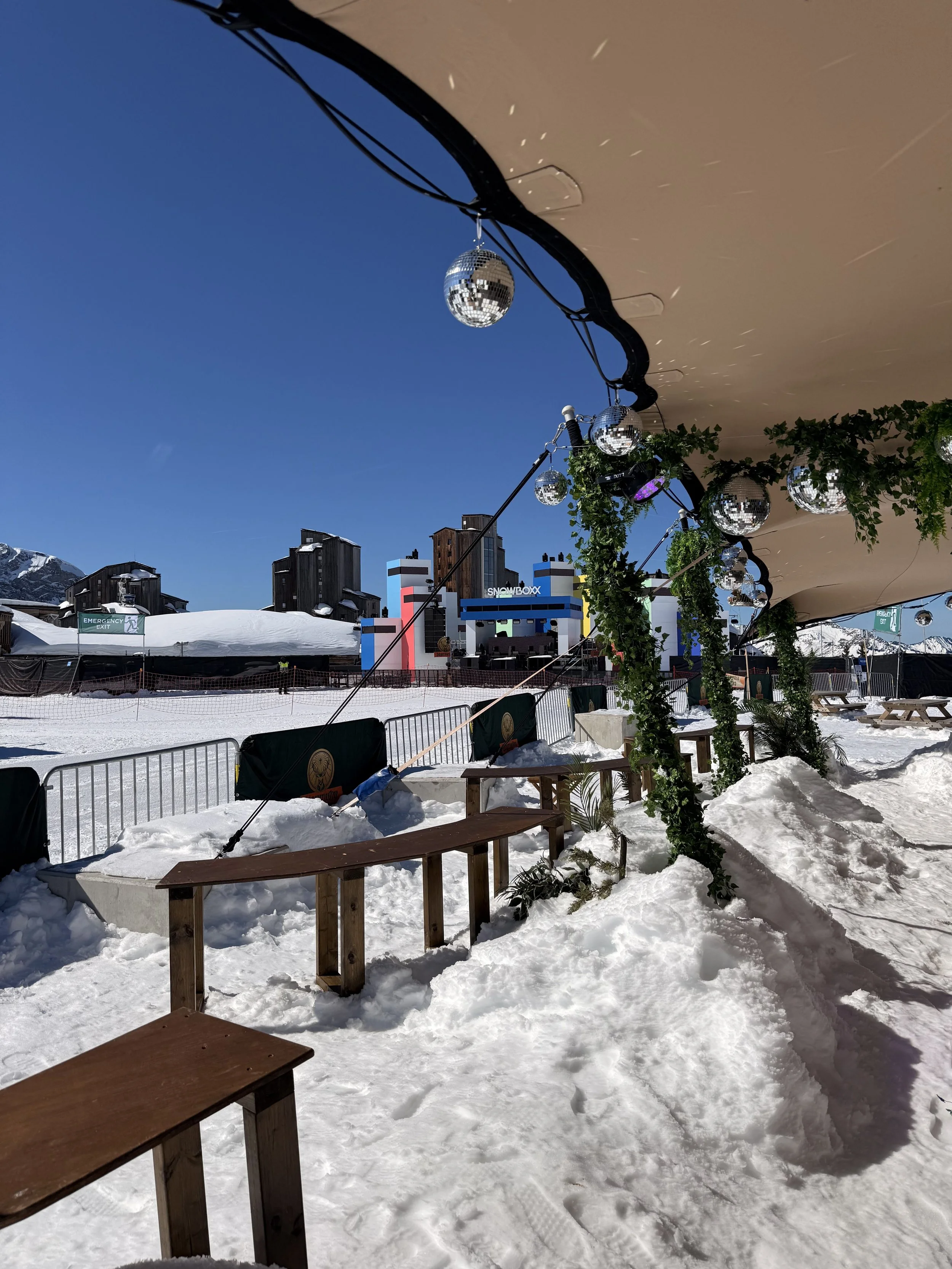 Outdoor winter scene with snow-covered ground, wooden benches, and a decorated tent with disco balls and greenery, set against a clear blue sky and ski resort buildings in the background.