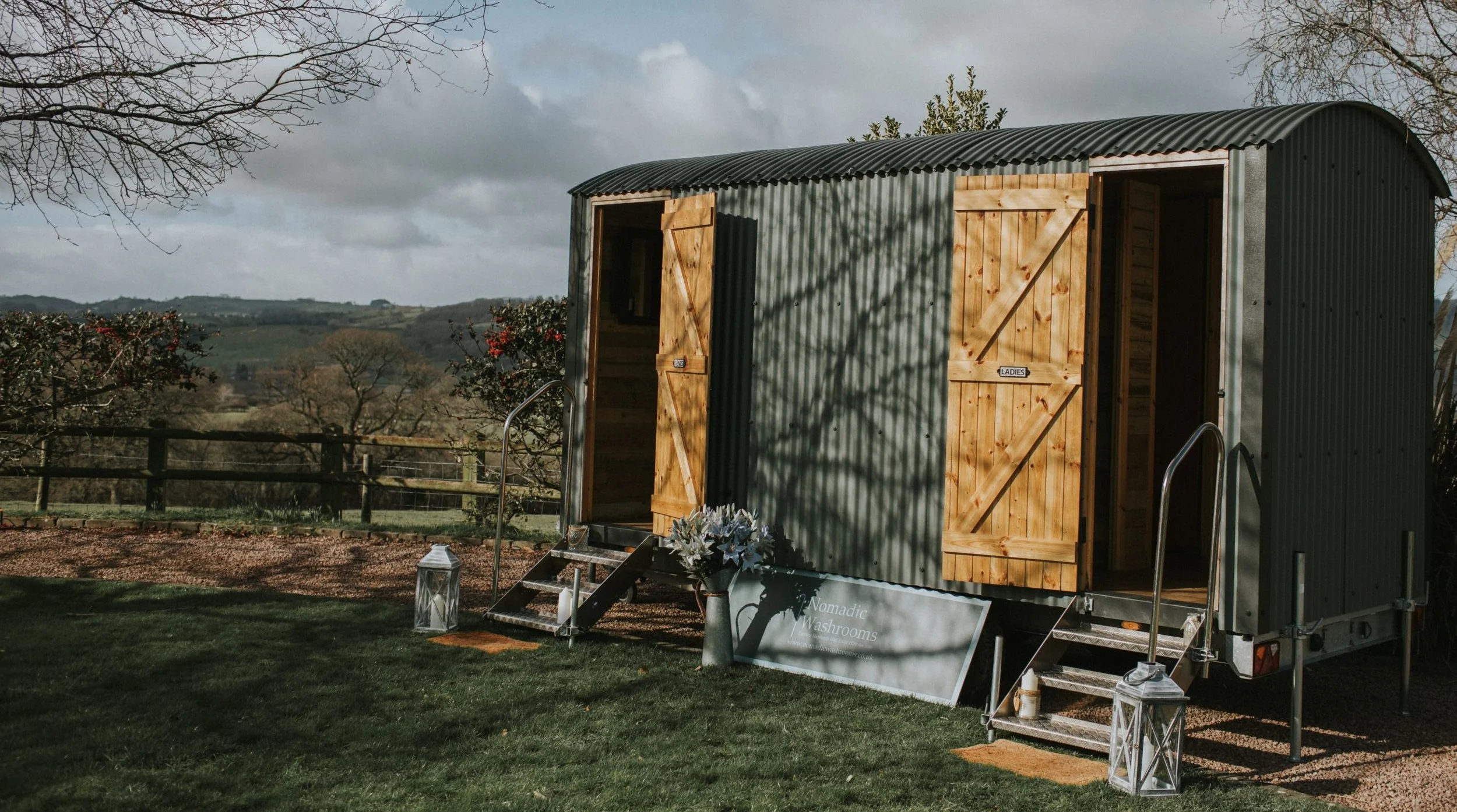 A tiny house on wheels with open wooden doors labeled 'Ladies' and 'Gents', set outdoors on a grassy area with a scenic countryside view, two lanterns, and a sign reading 'Nomadic Washrooms'.
