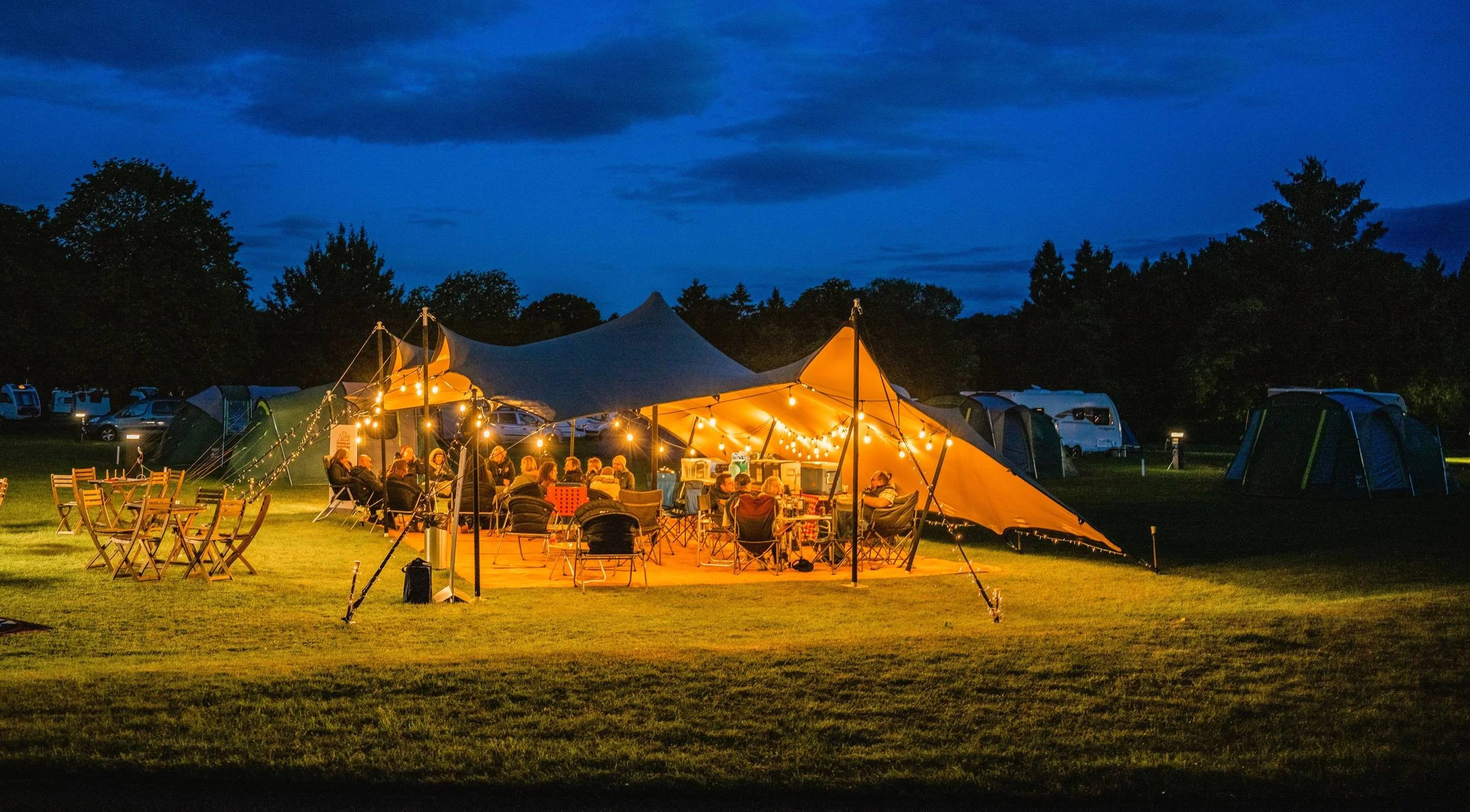 10 x 7 m stretch tent lit with festoon lights at dusk, set up for an evening party on a Cotswolds campsite