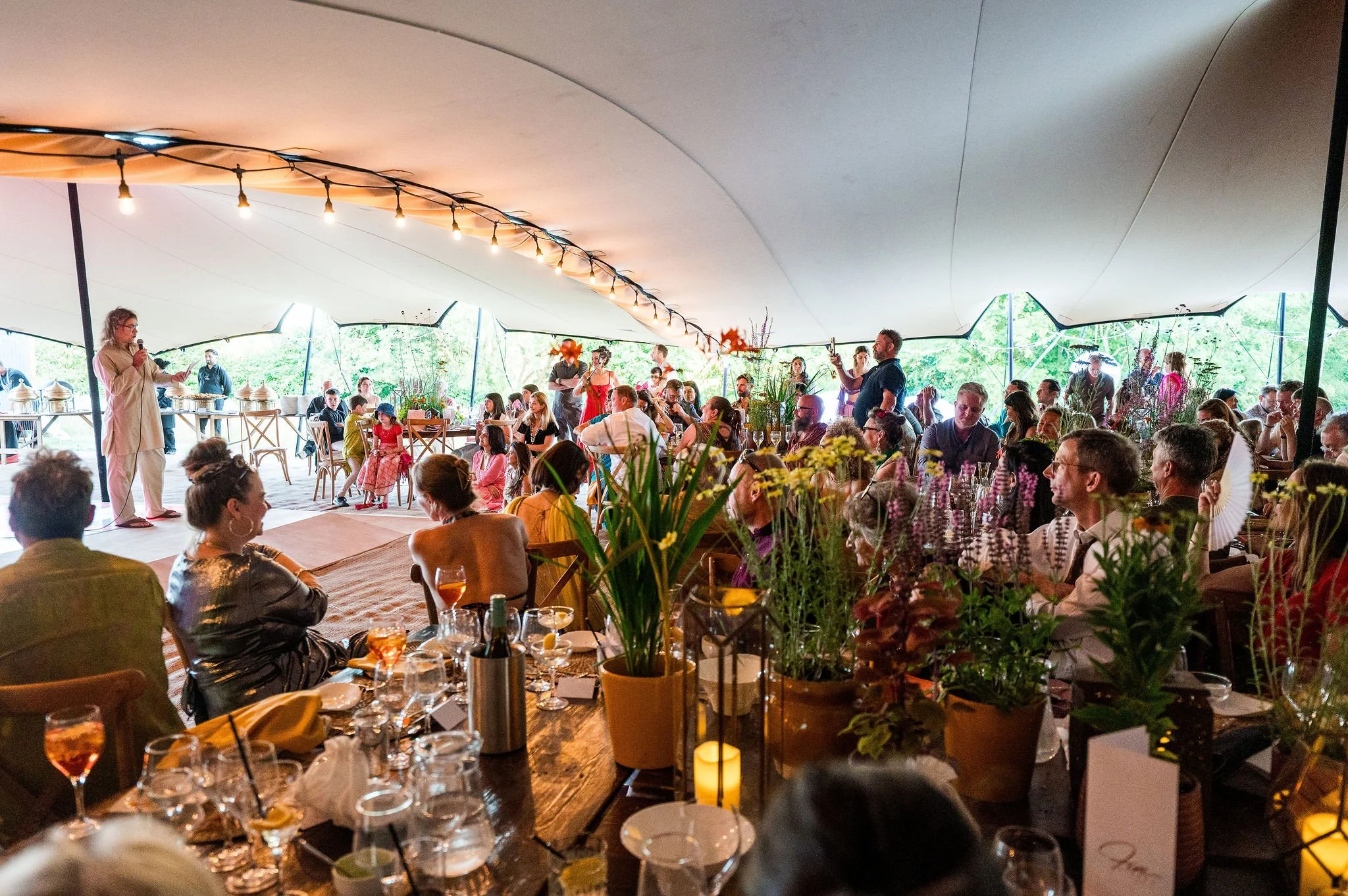 Guests seated at long tables with plants and candles inside a stretch tent lit by string lights