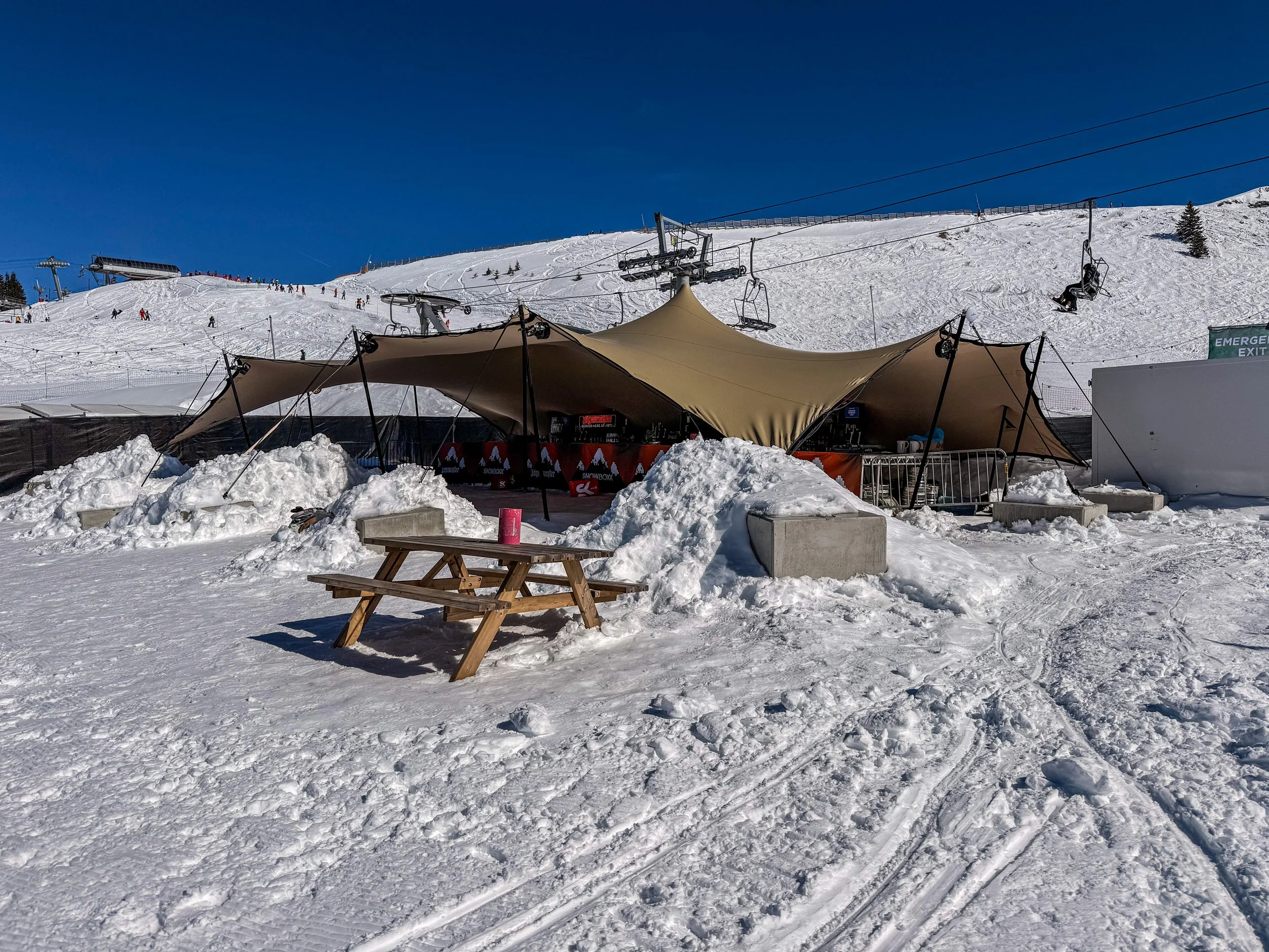 A ski resort with a snow-covered area, ski lifts, and a large beige canopy tent. There is a picnic table with a pink cup, snow piles, and ski tracks in the foreground, with skiers and snowboarders on the snowy slope in the background under a clear bl