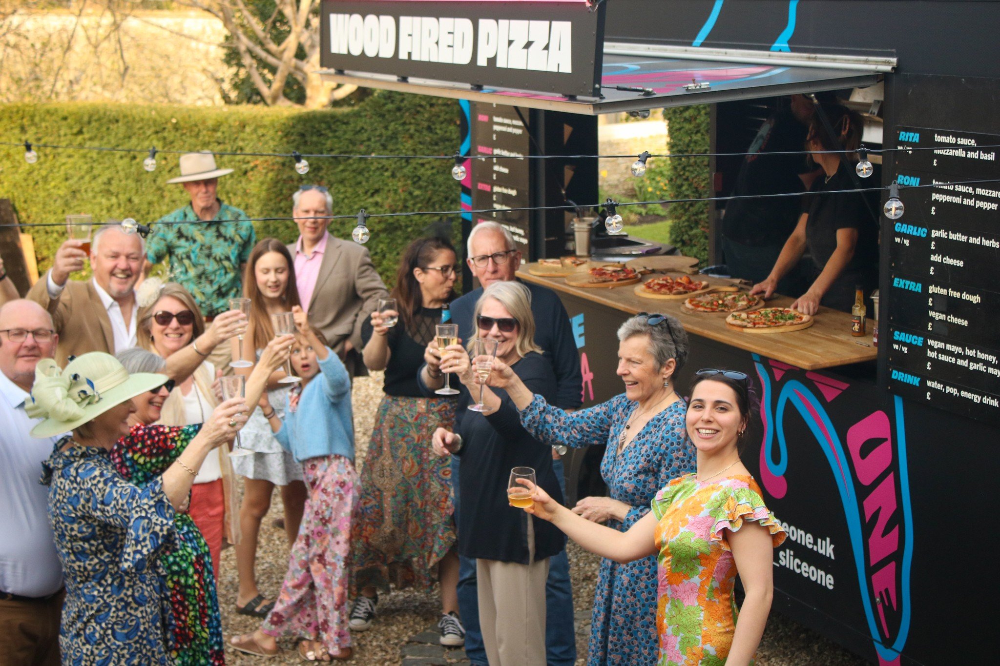 Group of people smiling and raising glasses in celebration in front of a pizza food truck at an outdoor event.
