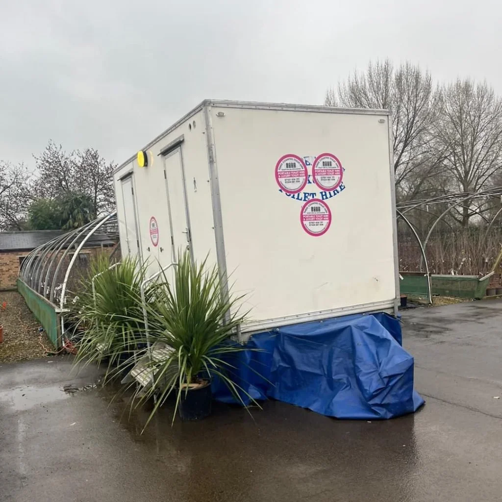 A small white portable building with a blue tarp covering part of the bottom and three pink and white stickers on the front, placed on a wet pavement outside. There are trees without leaves and a greenhouse structure nearby.