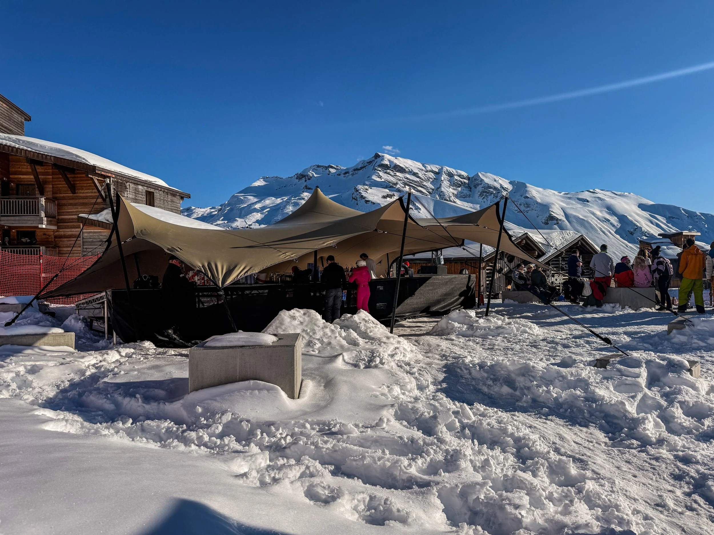 Snow-covered outdoor area with a large beige canopy tent and people gathered, wooden buildings on either side, snowy mountains in the background, and bright blue sky.