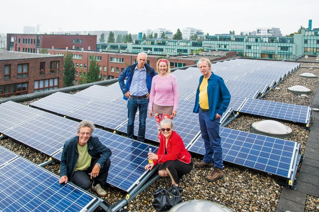 Five people, three men and two women, on a rooftop with solar panels, in front of a cityscape with modern apartment buildings.