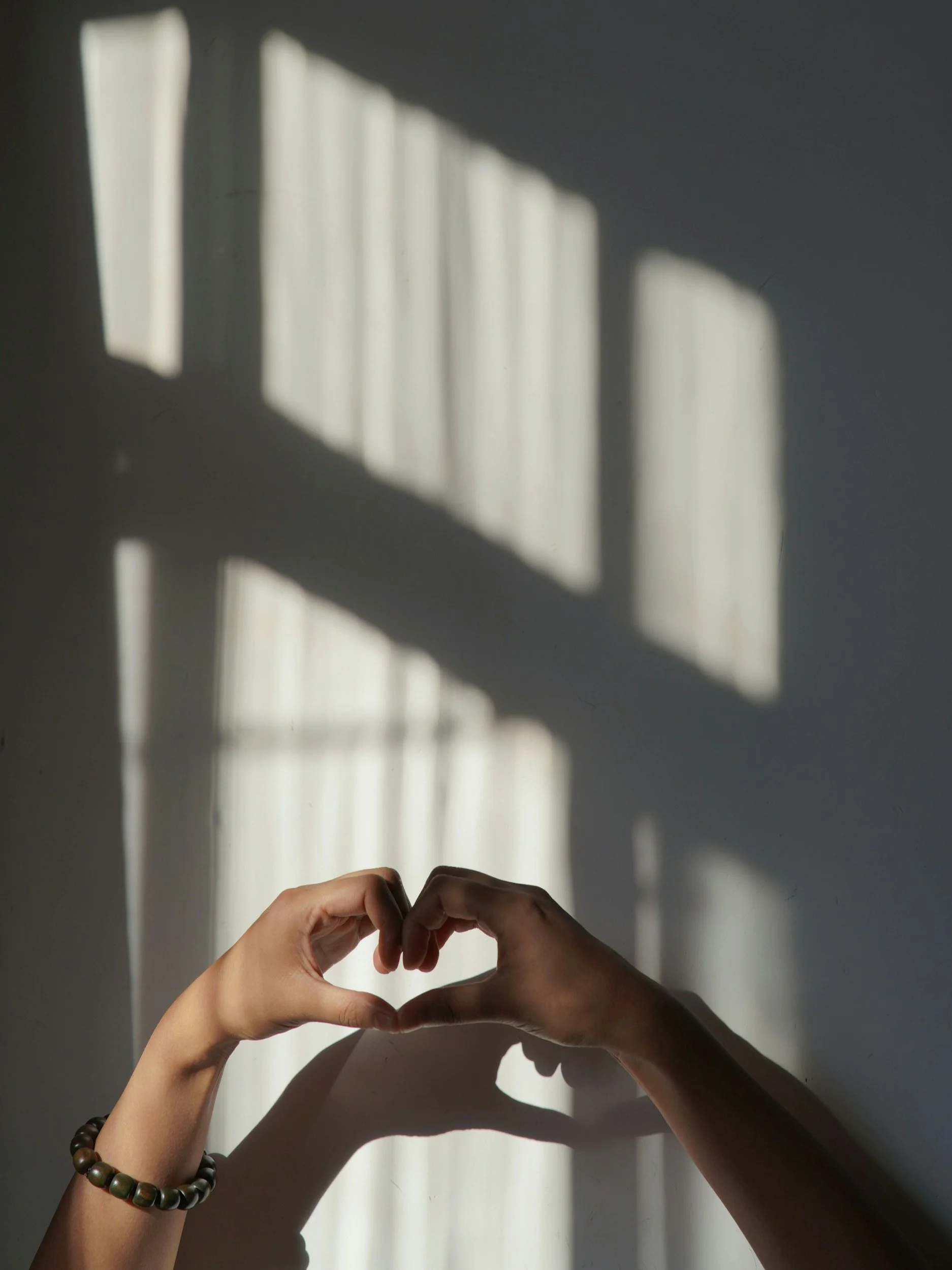 Hands forming a heart shape in front of a wall with window light shadows.